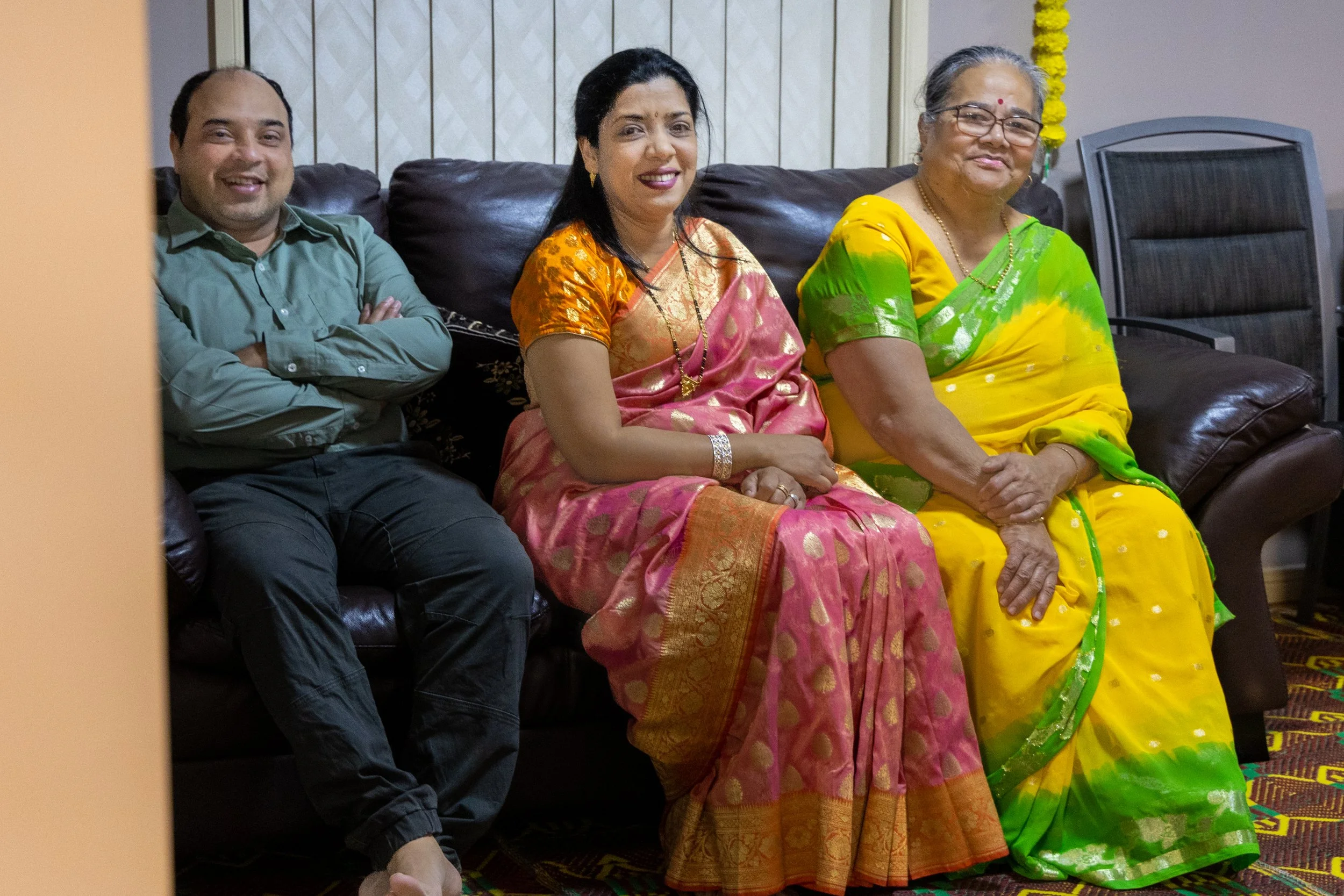 Three people sitting on a black leather sofa, smiling. A man on the left, wearing a green shirt and black pants, with arms crossed. A woman in the middle wearing a pink and gold sari with jewelry. An older woman on the right wearing a yellow and gree