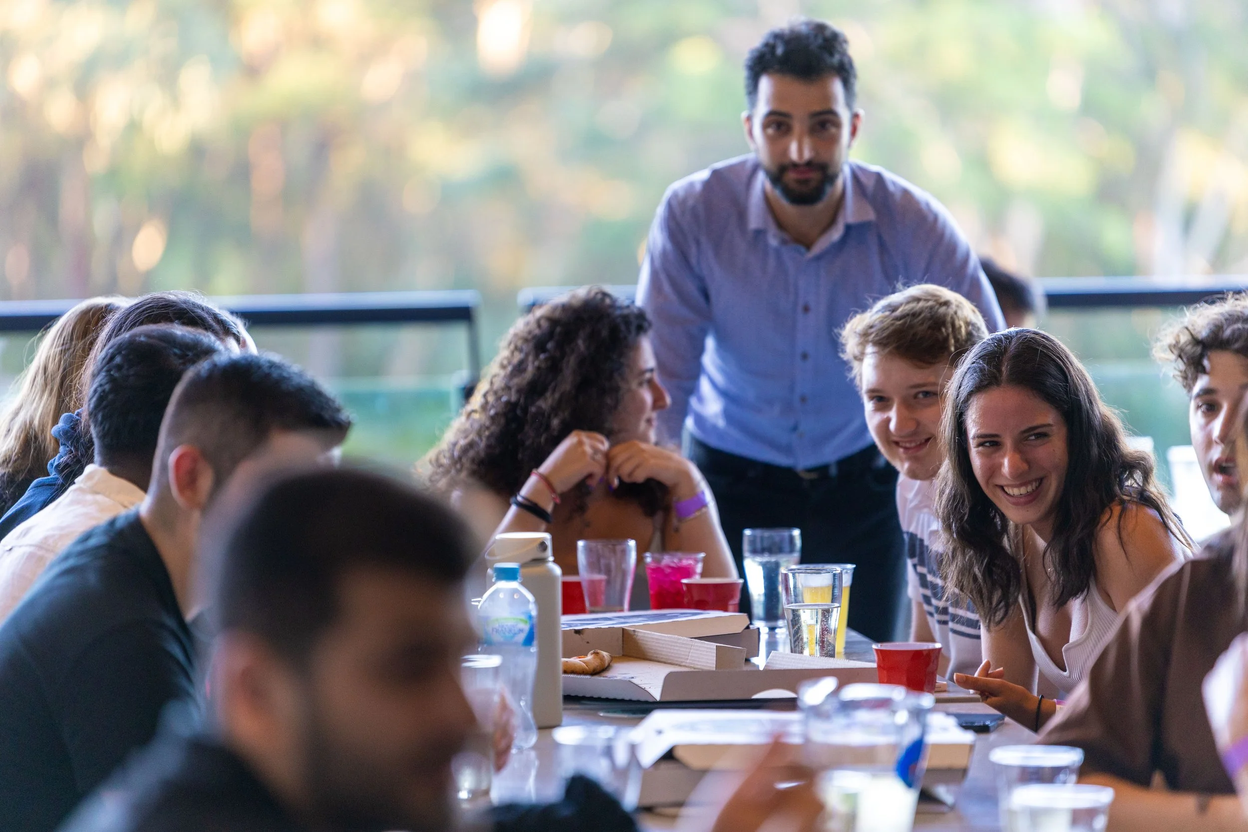 A group of diverse young people gathered around a table outdoors, smiling and listening to a man standing and speaking.