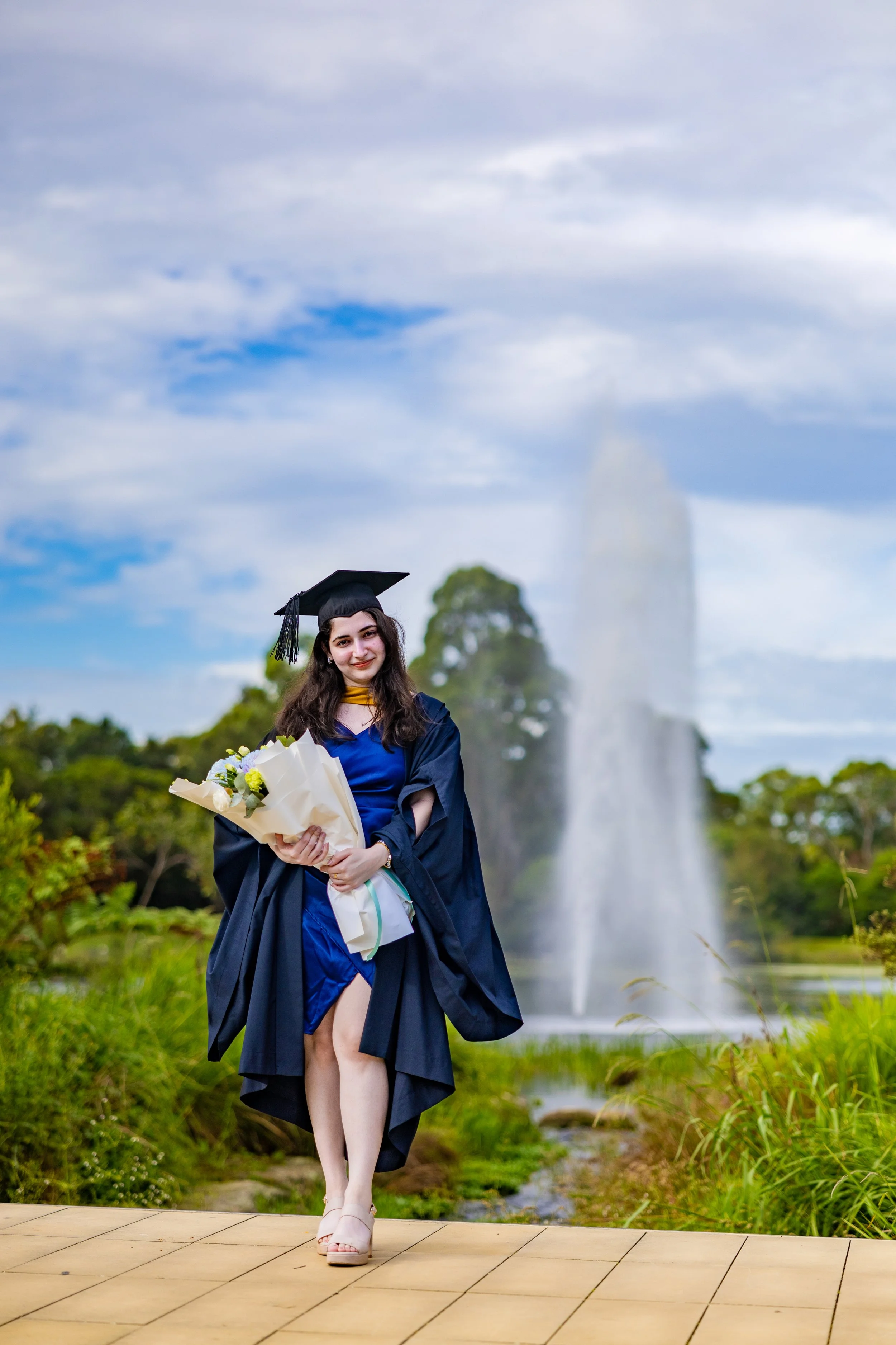 A young woman in a blue graduation gown and cap holding a bouquet of flowers, standing on a wooden walkway outdoors with a fountain and trees in the background.