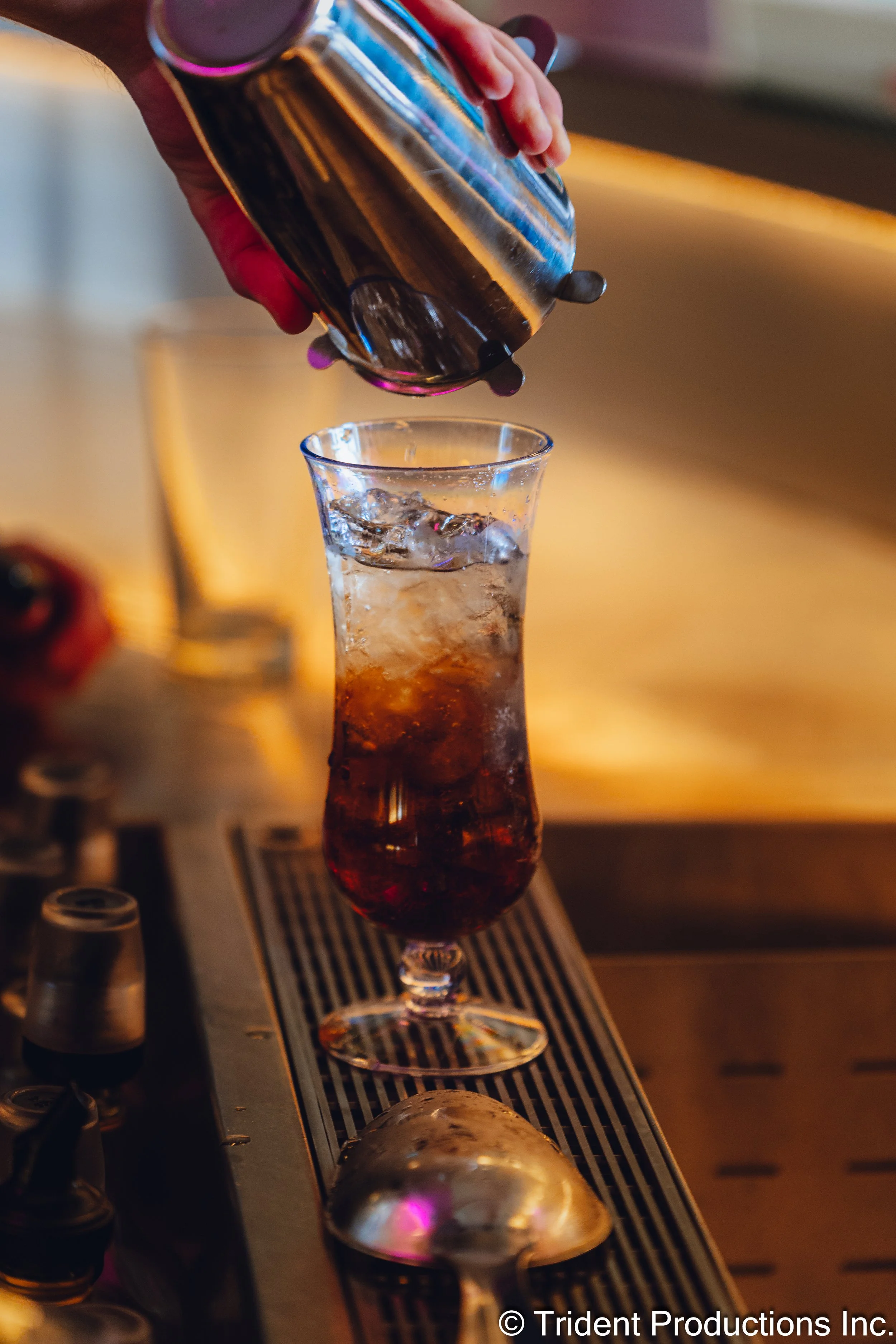 A hand pouring a dark soda from a can into a tall glass with ice on a bar counter.