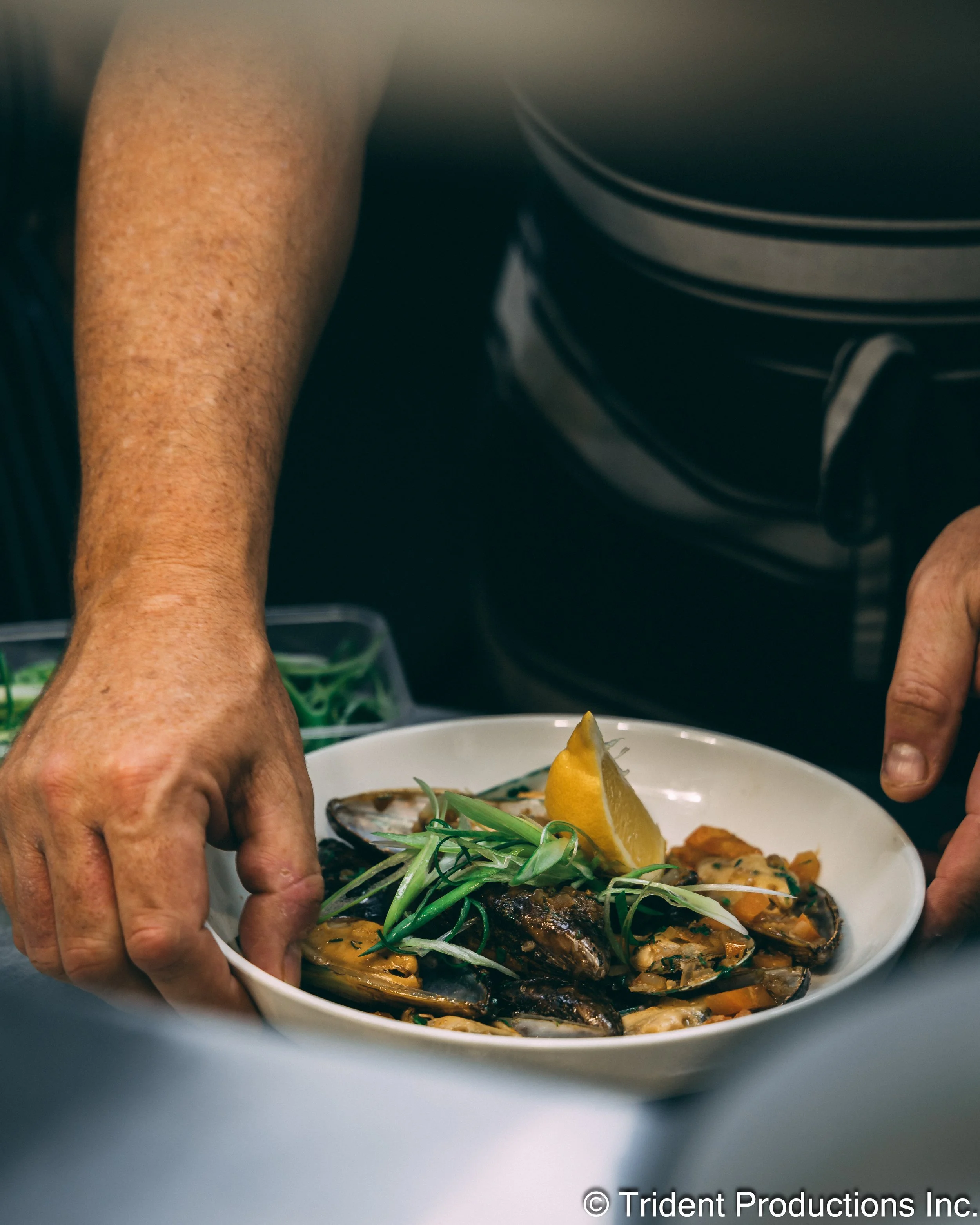 A person is placing a dish of cooked mussels garnished with green herbs and lemon wedges on a table.