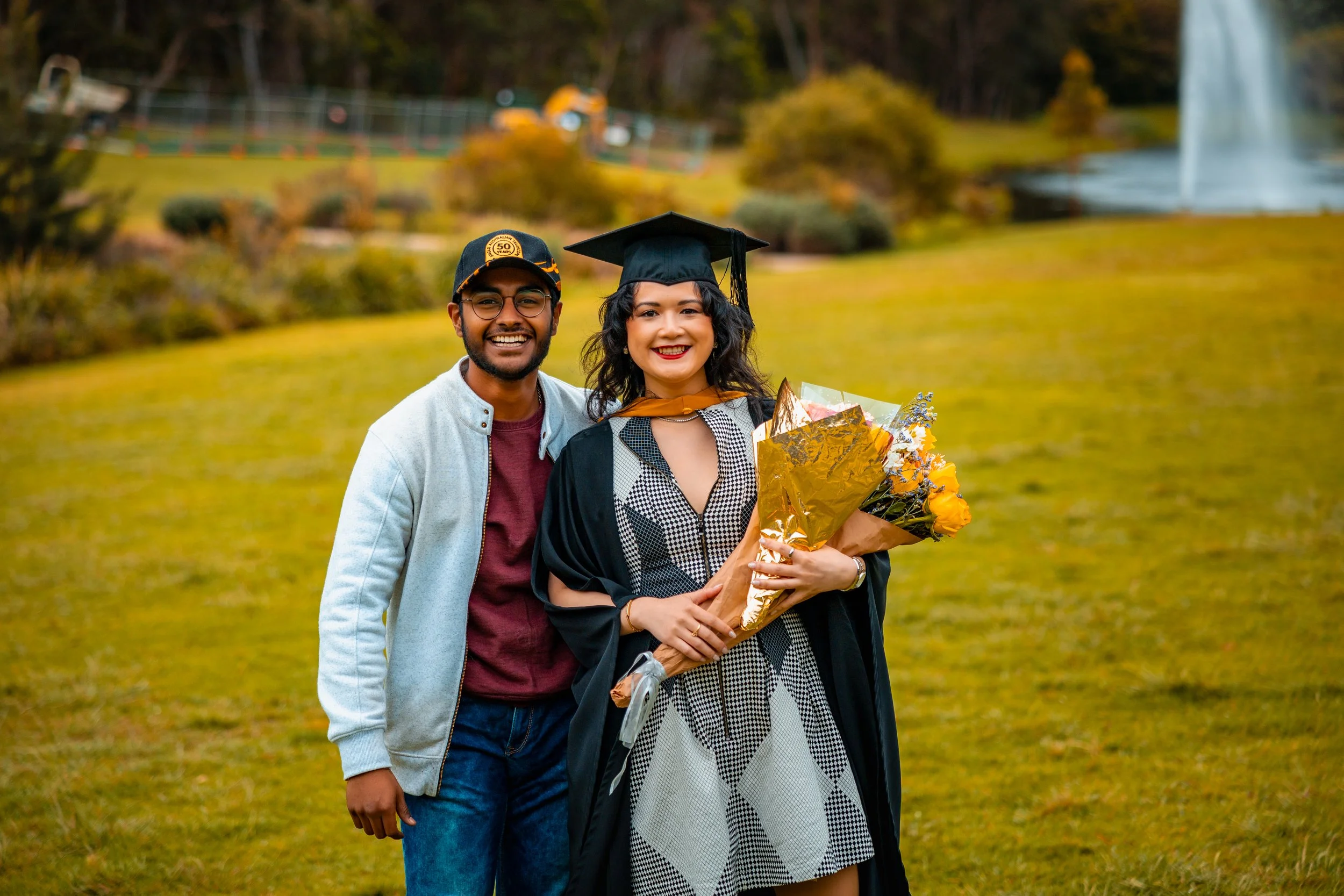 Happy woman in graduation gown and cap holding a bouquet of flowers, standing outdoors with a smiling man beside her in a park with a fountain in the background.