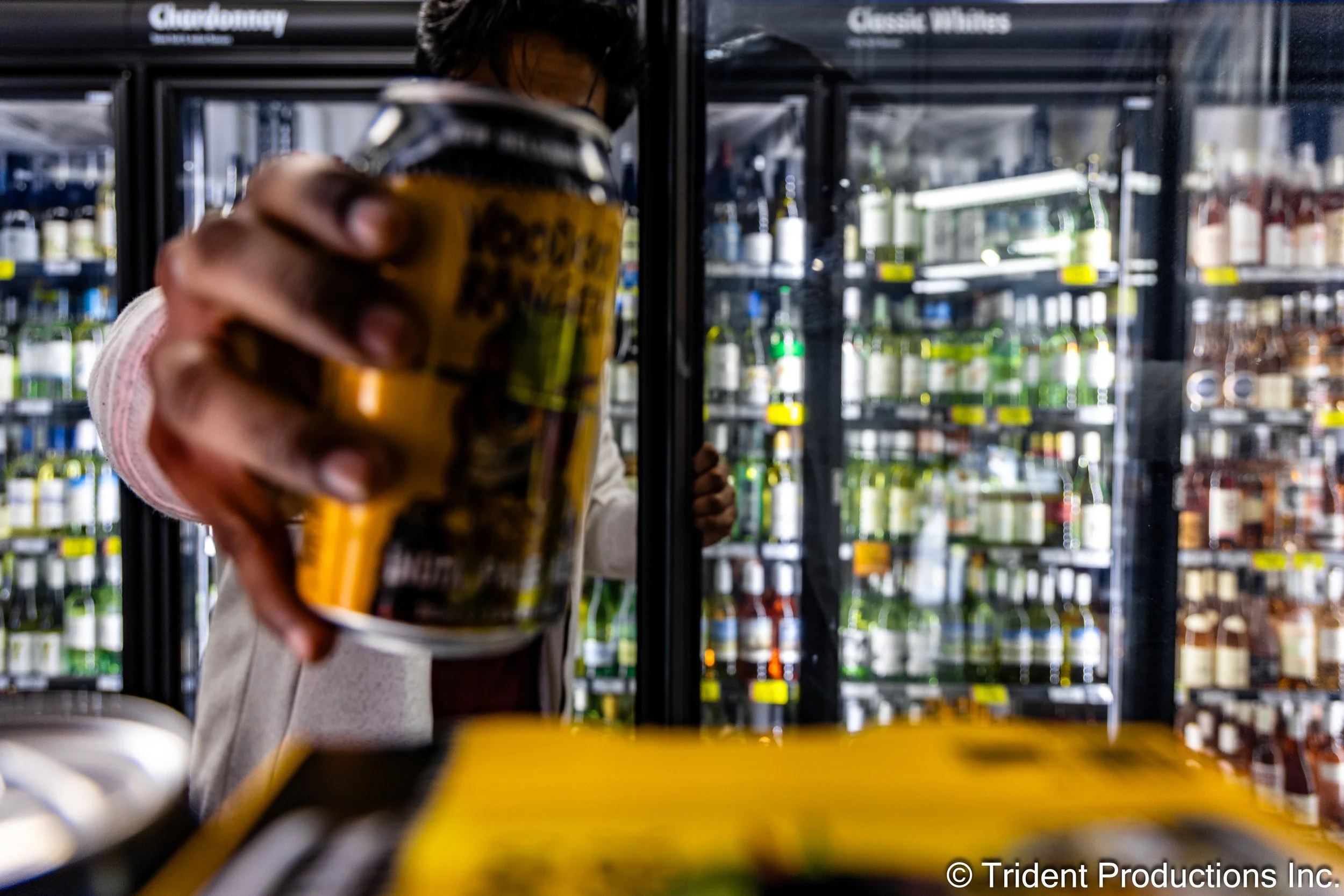 Person holding a can of beer in front of a glass door refrigerator stocked with various bottles, in a store.