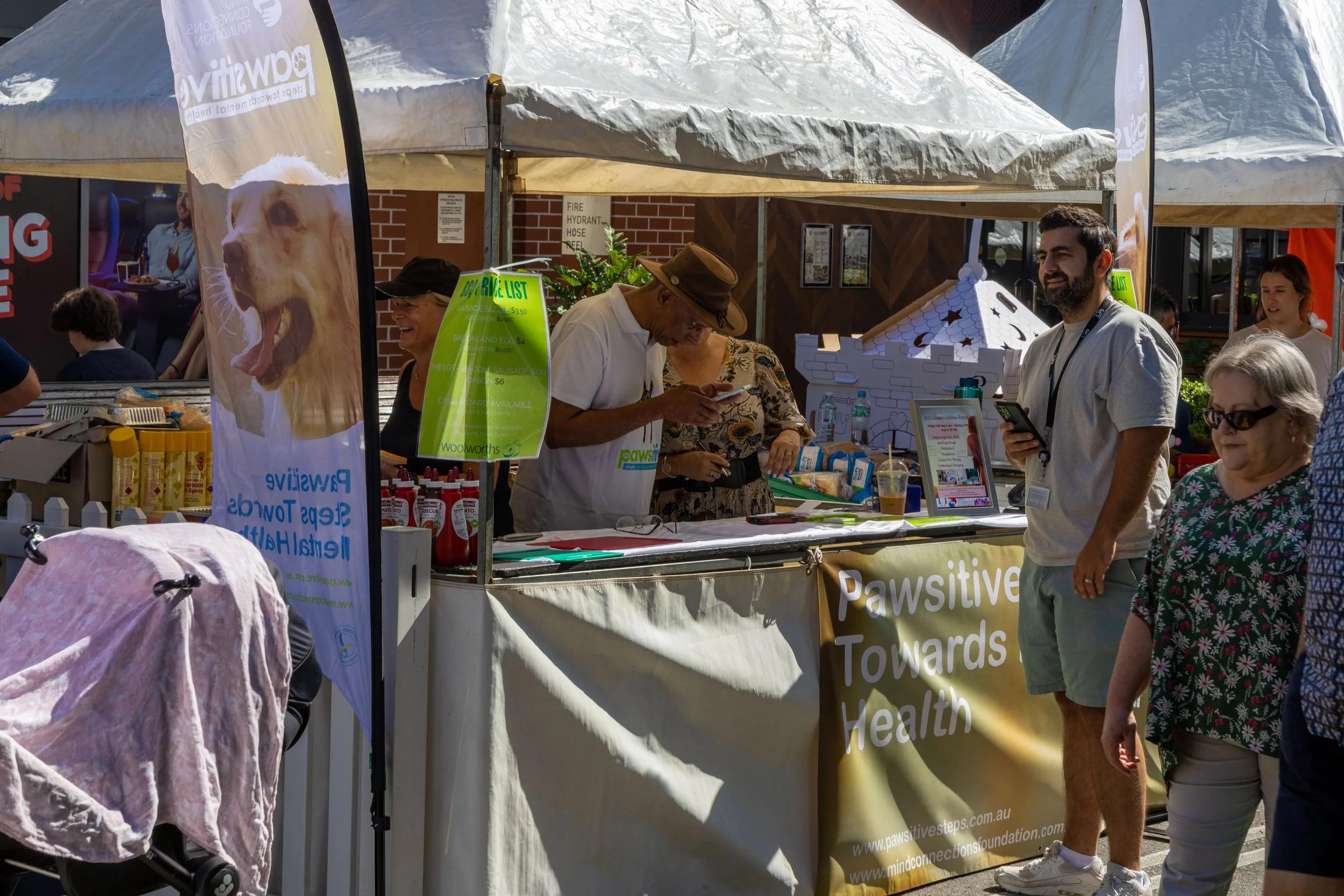 Outdoor event with a booth labeled 'Pawsitive Toward Health,' people socializing, and a sign offering a price list for items such as sausages and salads. The booth has a banner with a large picture of a yawning dog.