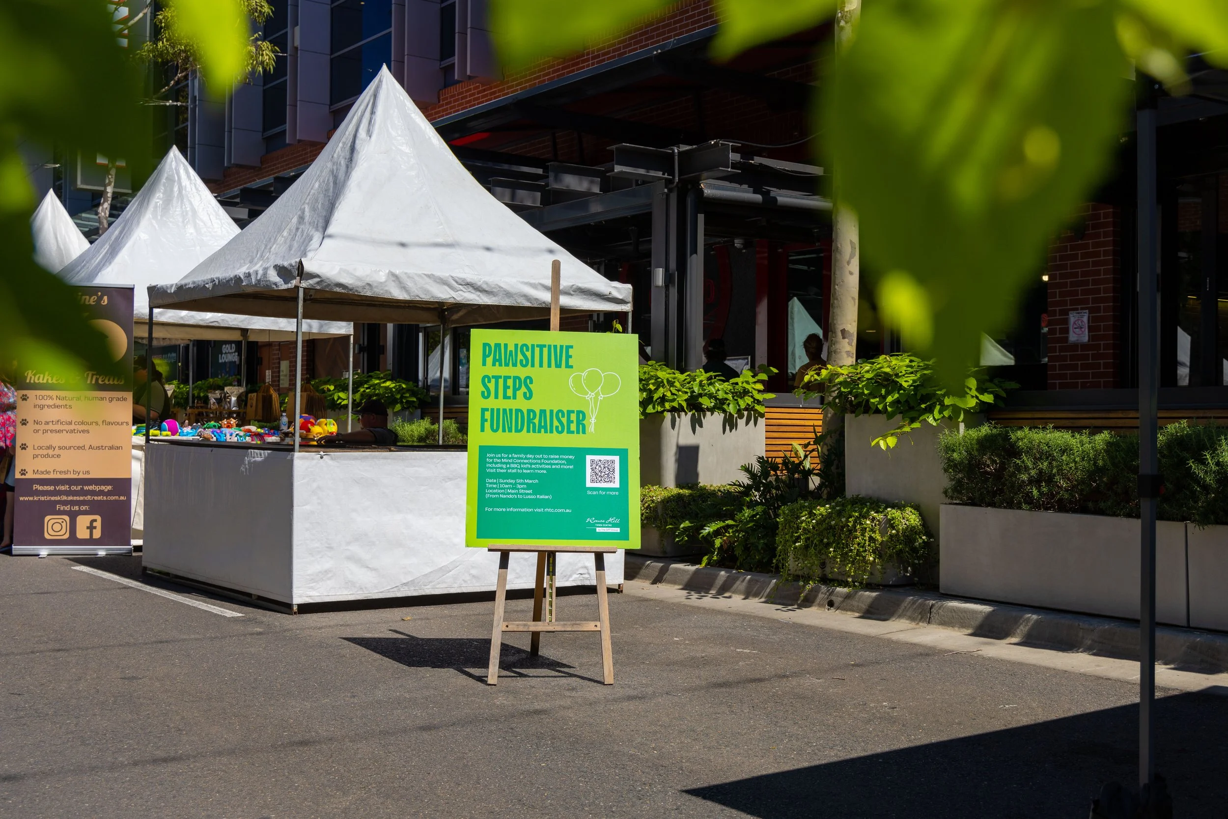 An outdoor market stall with a white canopy, displaying colorful toys, and a green sign that reads "Pawsitive Steps Fundraiser" with a QR code, set against a building with large windows and potted plants.