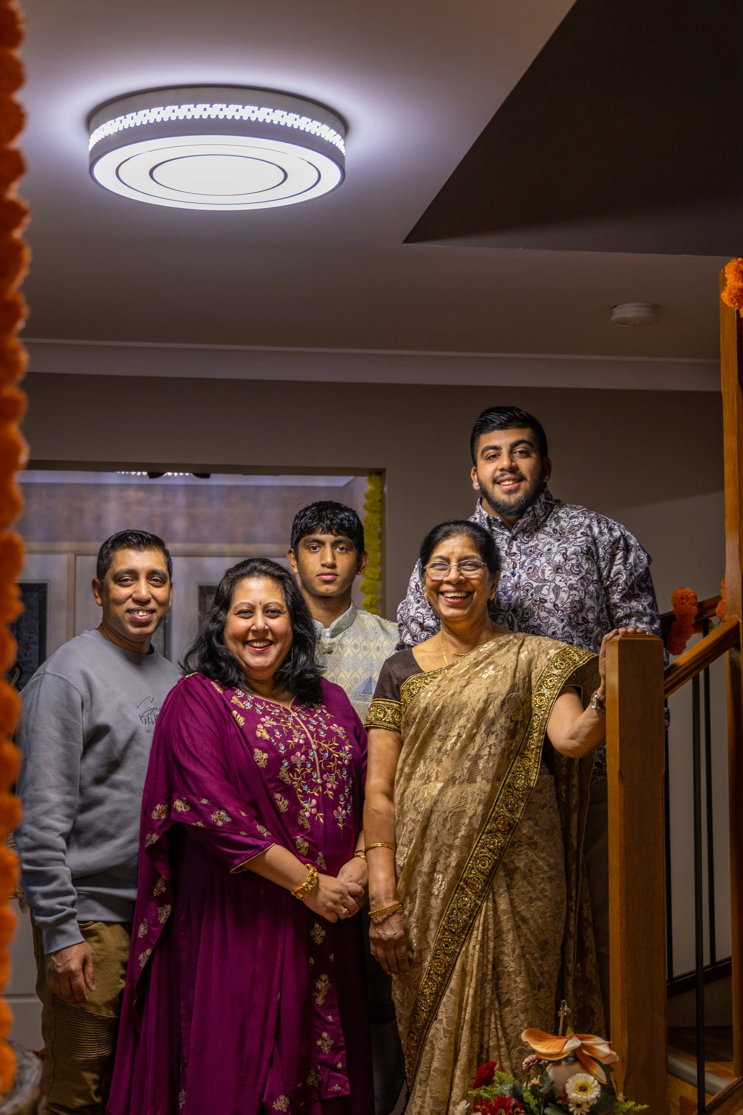 A group of six people posing together indoors, smiling for the camera, with warm lighting and decorative flowers in the foreground.