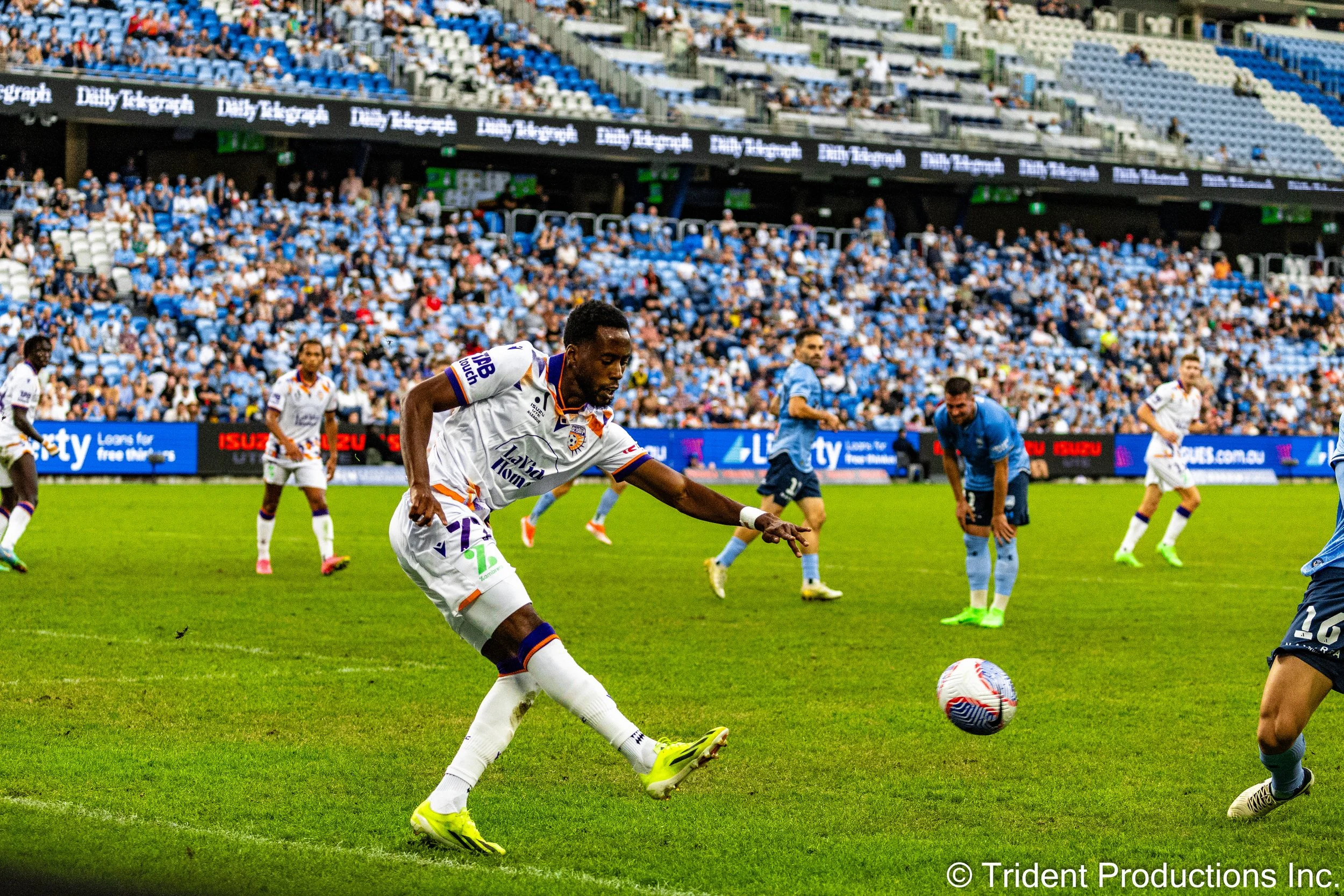 A soccer player in a white uniform kicking a soccer ball during a game, with other players and a stadium full of spectators in the background.