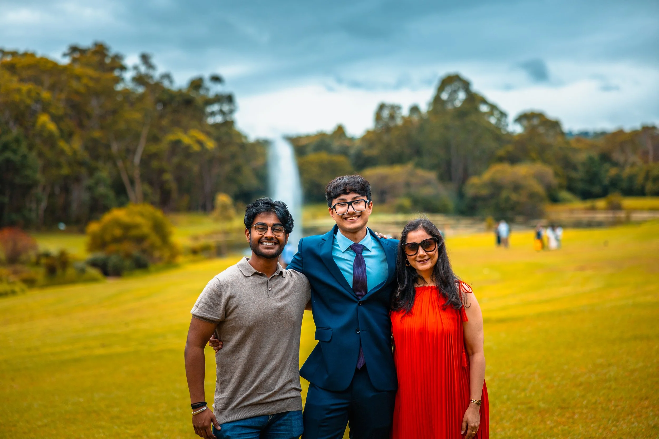 Three people smiling and standing together outdoors in front of a waterfall with trees and grassy field in the background.