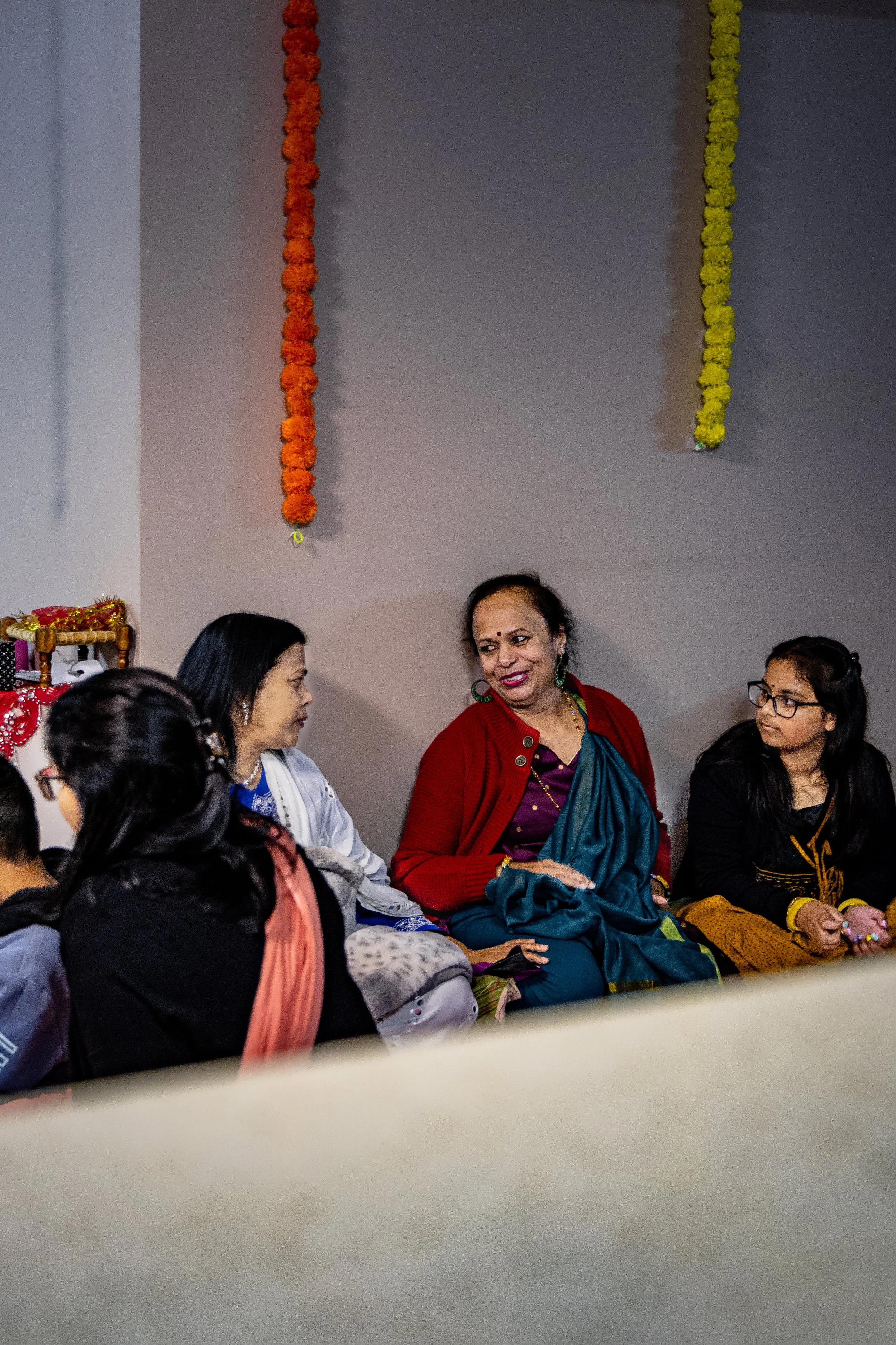 A group of women sitting on the floor and engaging in conversation, decorated with orange and yellow flower garlands hanging on the wall behind them.