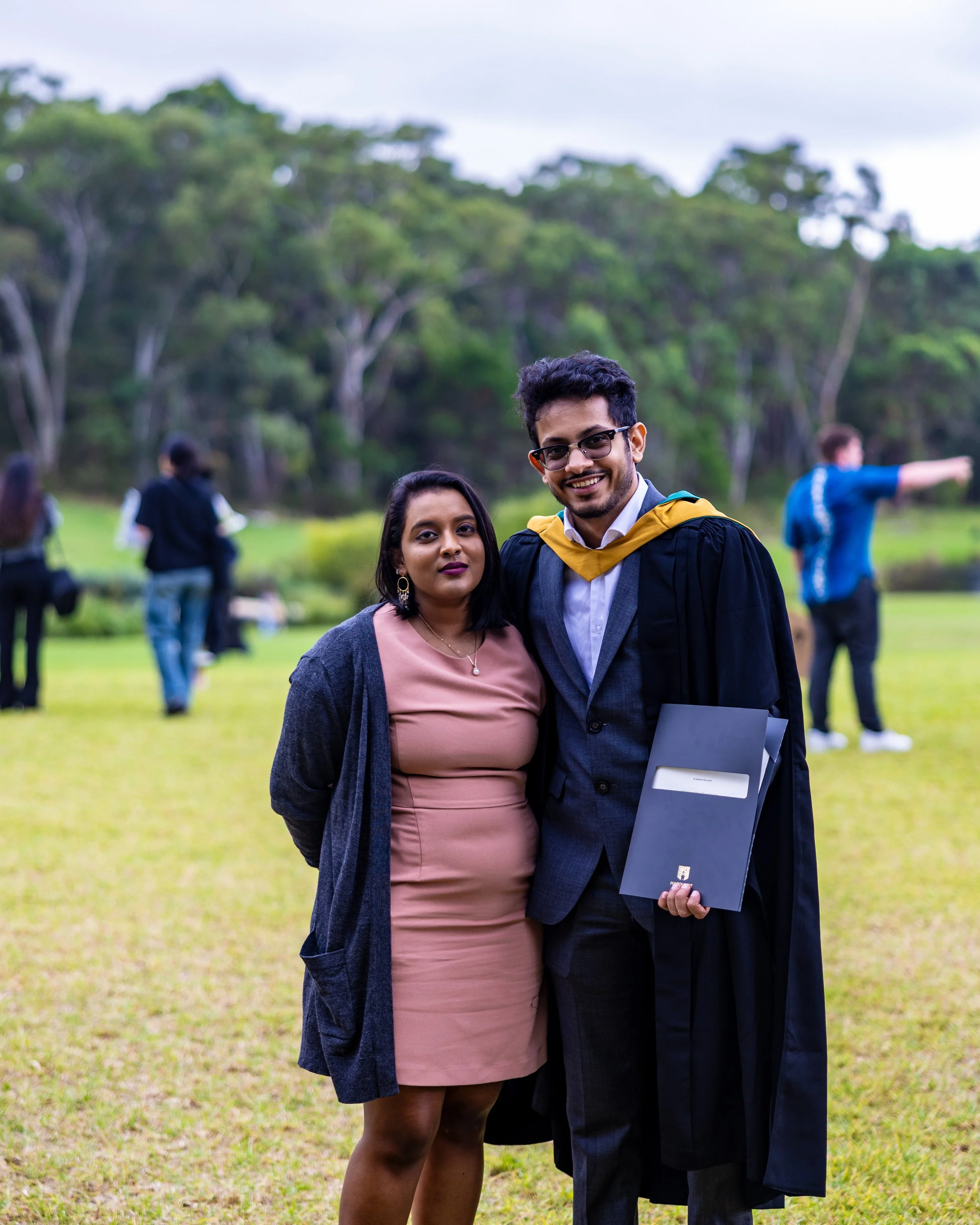 A graduate in a black gown and cap with a yellow hood standing next to a woman in a pink dress and dark cardigan outdoors with trees in the background, during a graduation celebration.