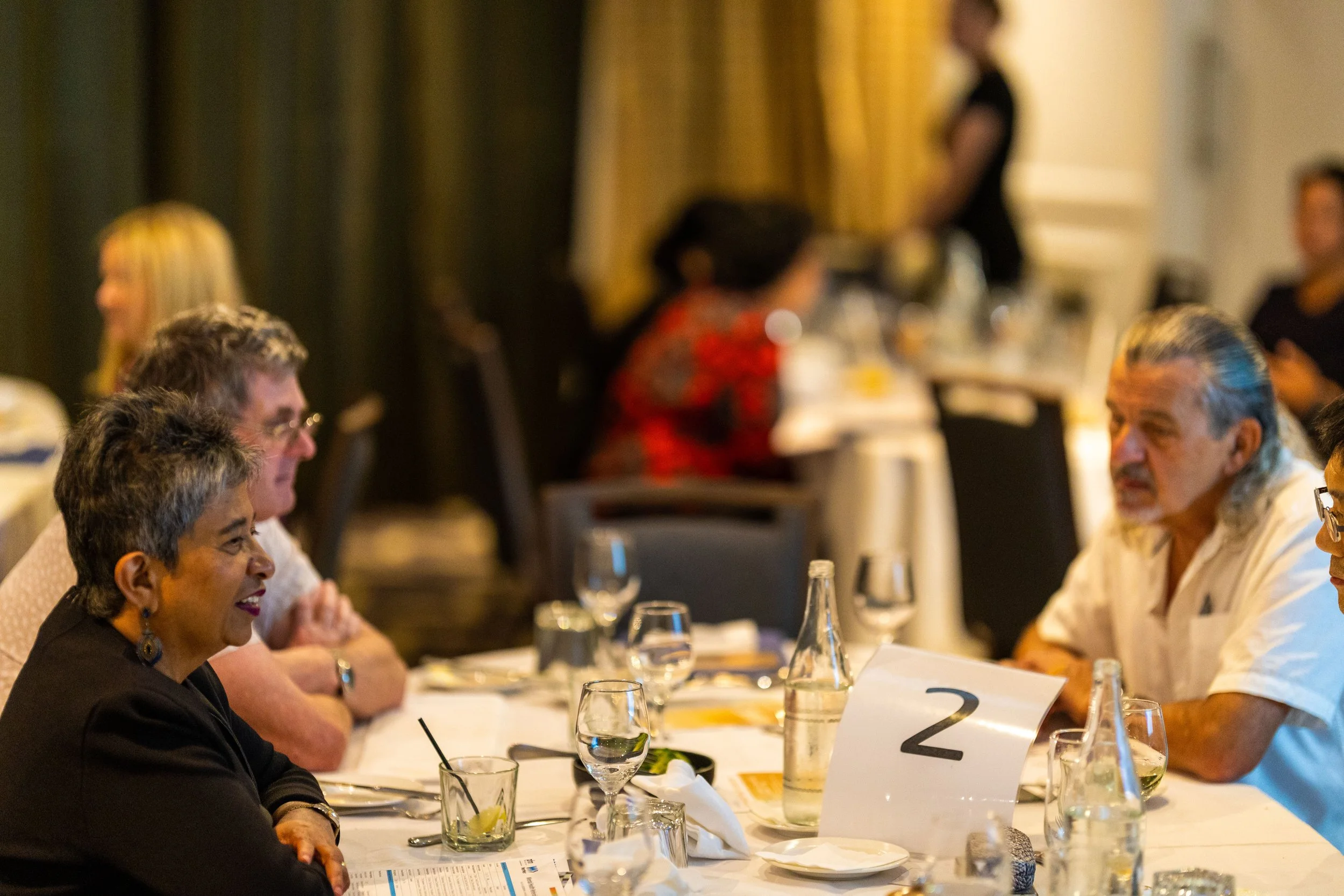 People sitting around a dinner table engaged in conversation, with glasses, drinks, and a table number '2' visible.