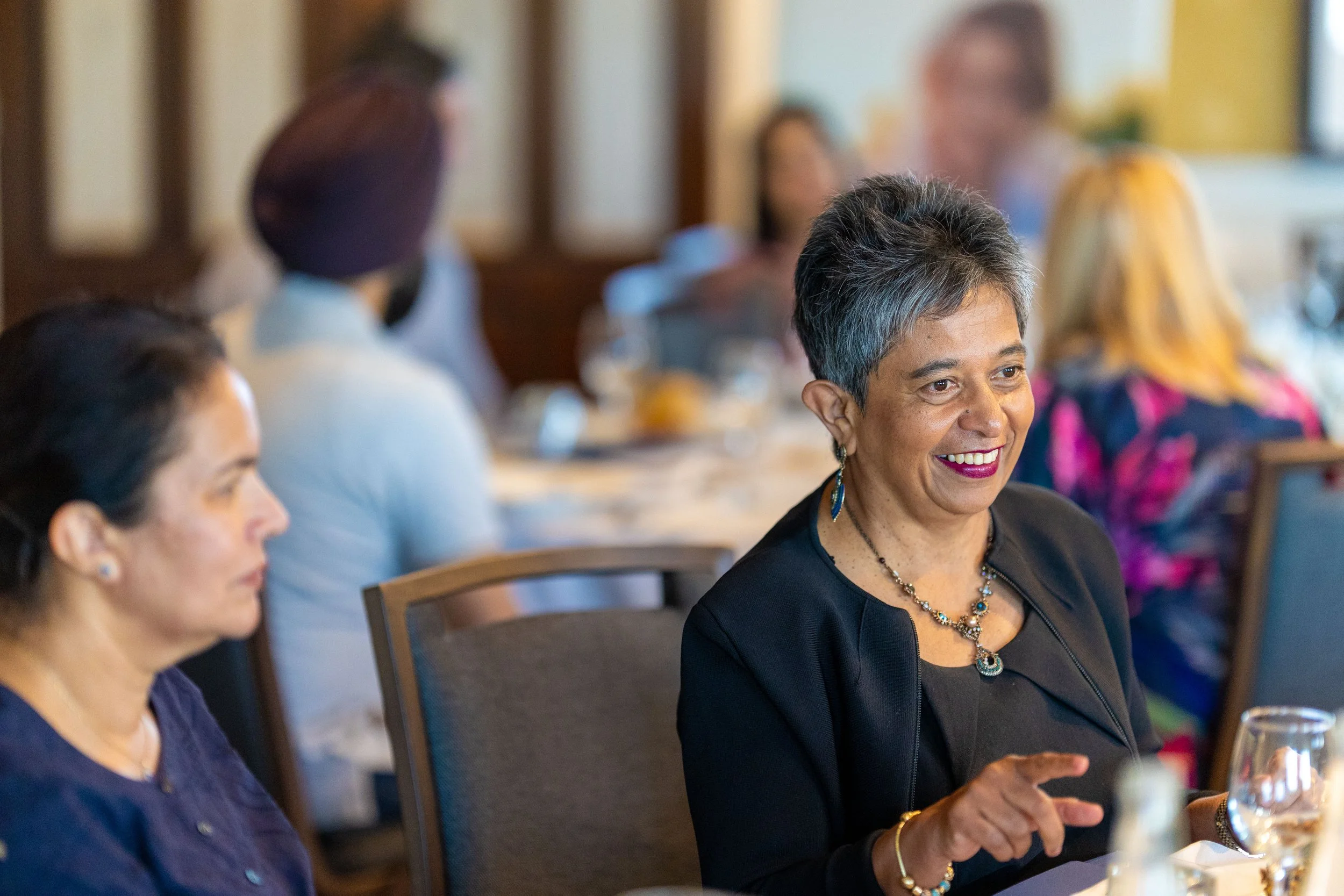 A group of women sitting around a dining table, with one woman in the foreground smiling and talking.