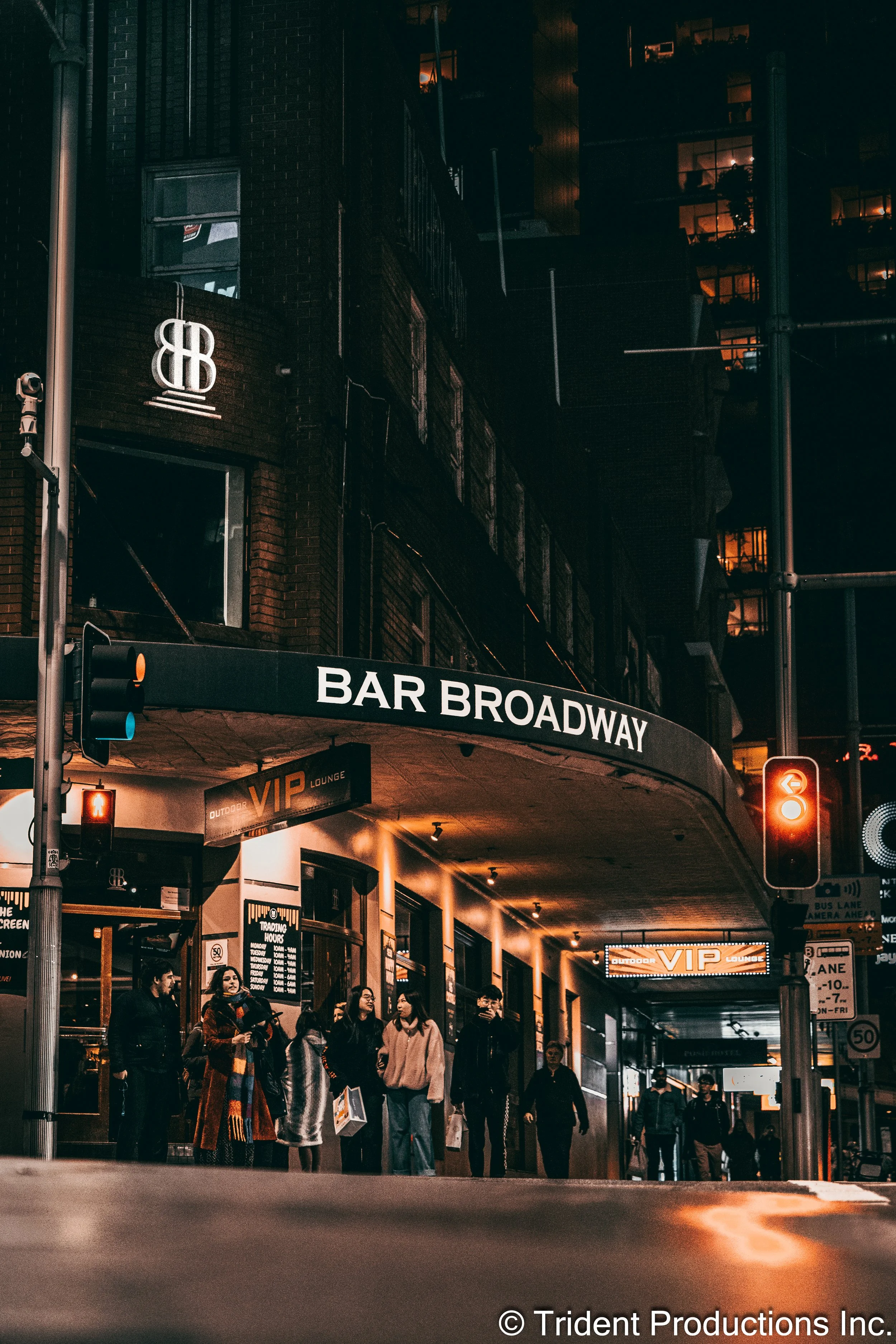 Nighttime scene outside Bar Broadway with people standing outside the entrance, illuminated signs, and buildings in the background.