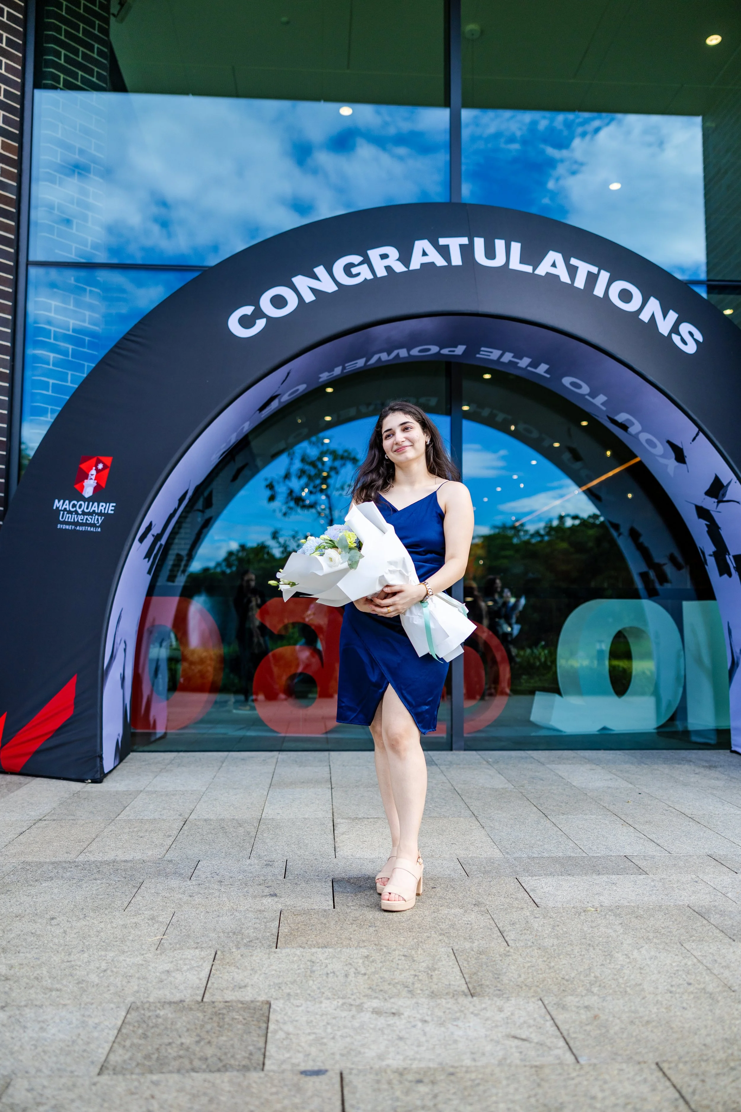 A young woman in a blue dress holding a bouquet of flowers standing outside a building with a banner that says, 'Congratulations,' celebrating her graduation at Macquarie University in Australia.