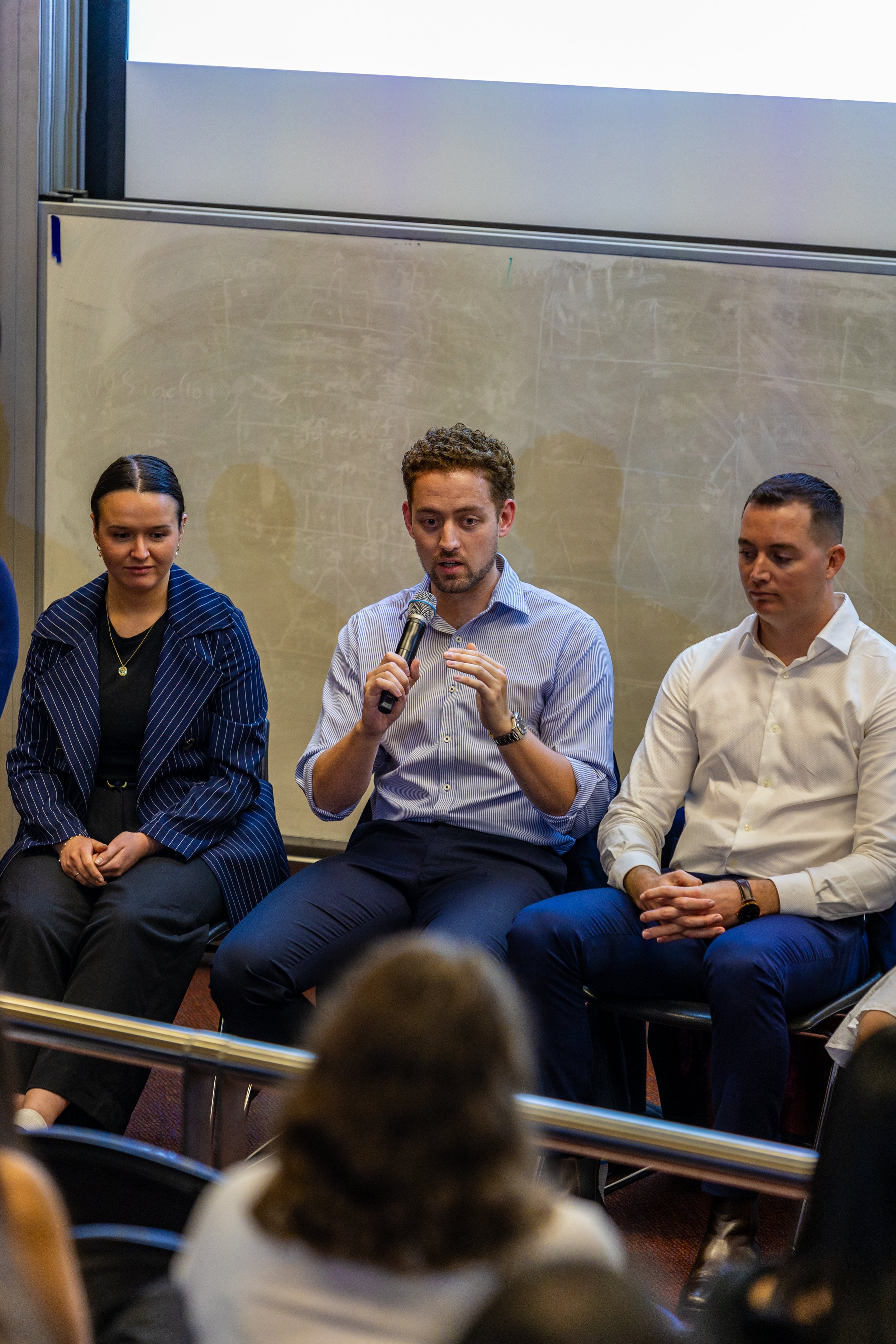 A panel discussion with three people seated on stage in front of an audience. The man in the middle is speaking into a microphone, while the woman on the left and the man on the right listen. The background features a chalkboard.