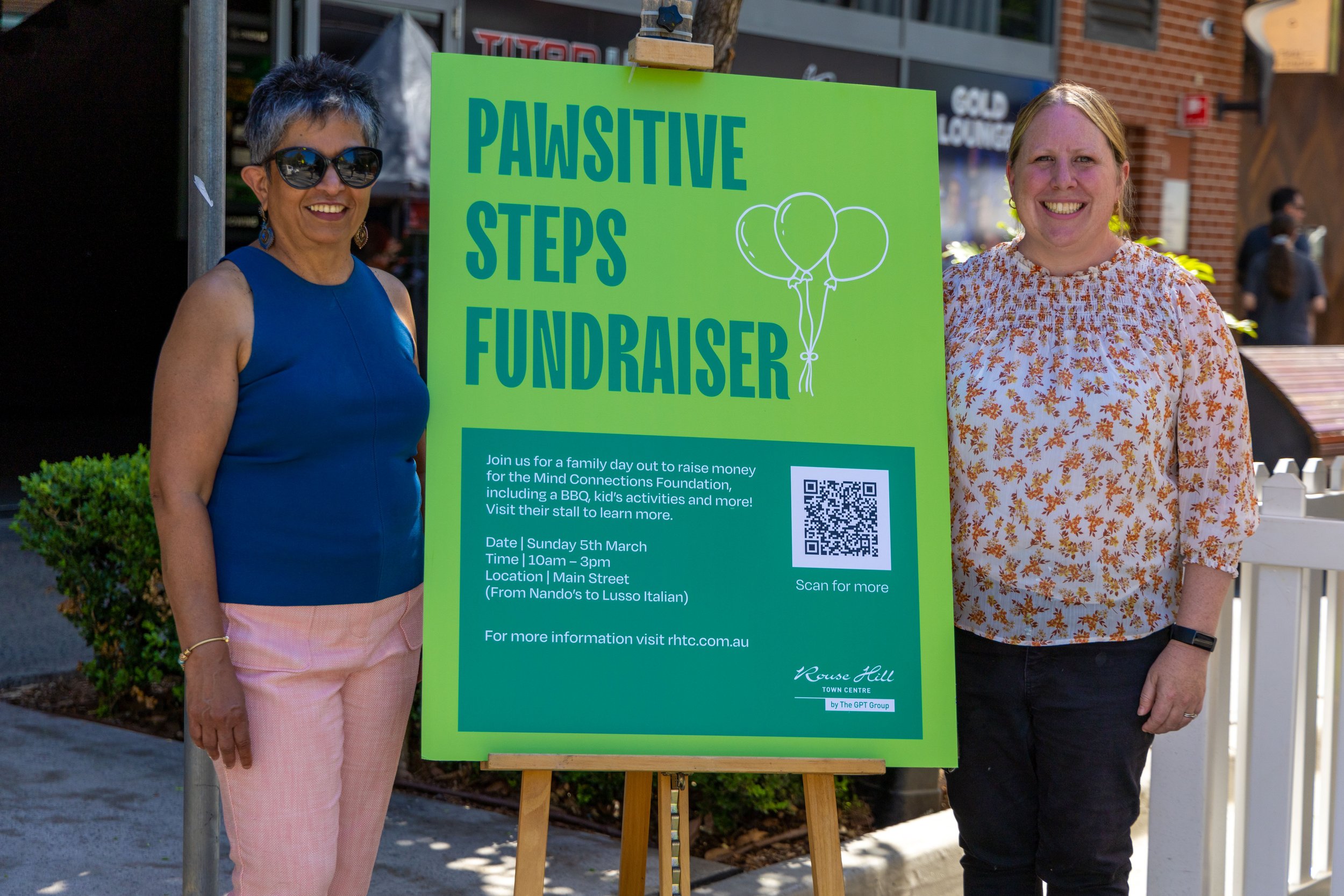 Two women standing outdoors next to a bright green sign that reads 'Pawsitive Steps Fundraiser' with balloons illustration, providing event details including date, time, location, and a QR code.