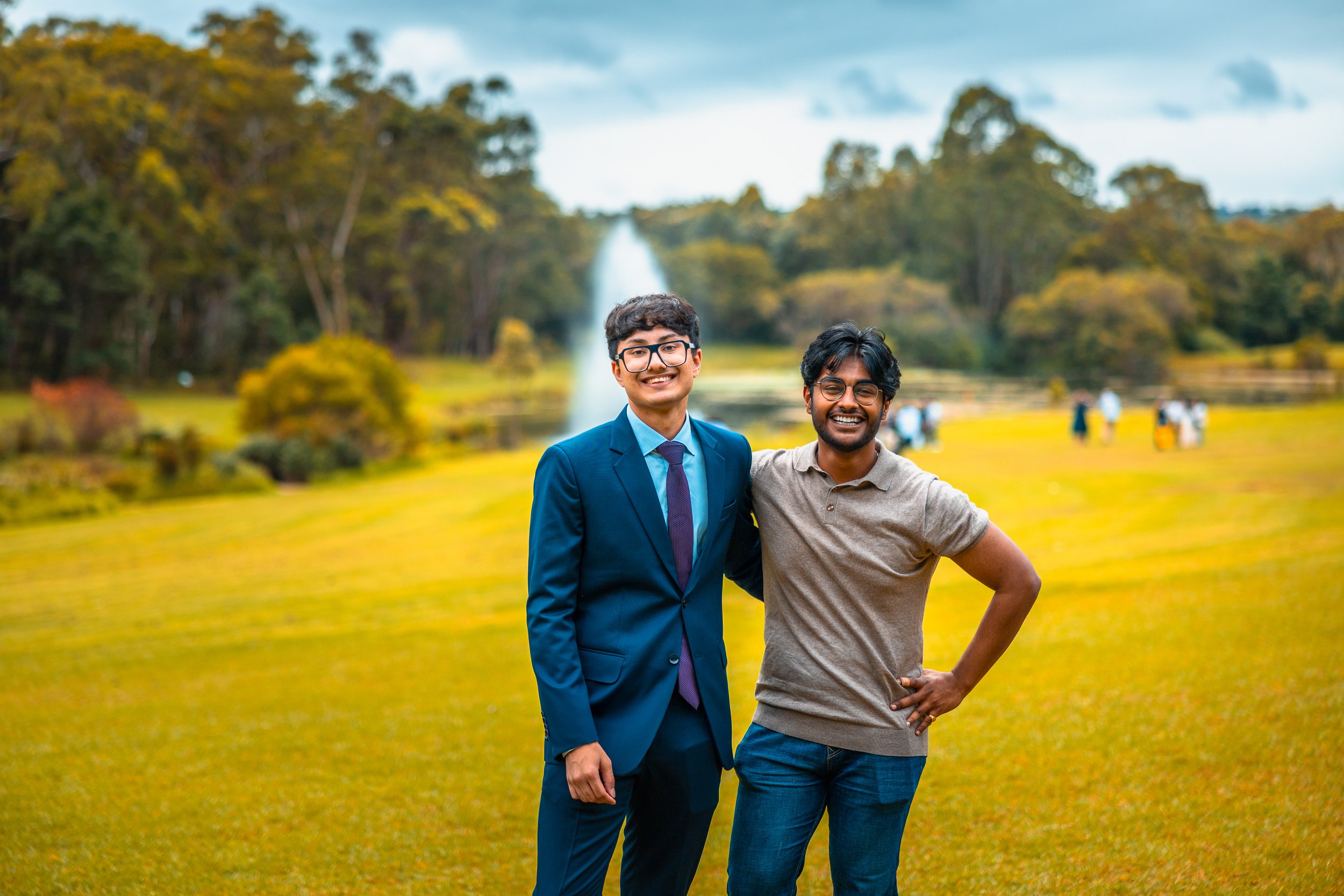 Two men smiling and standing outdoors on a grassy field near trees and a fountain, with other people in the background.