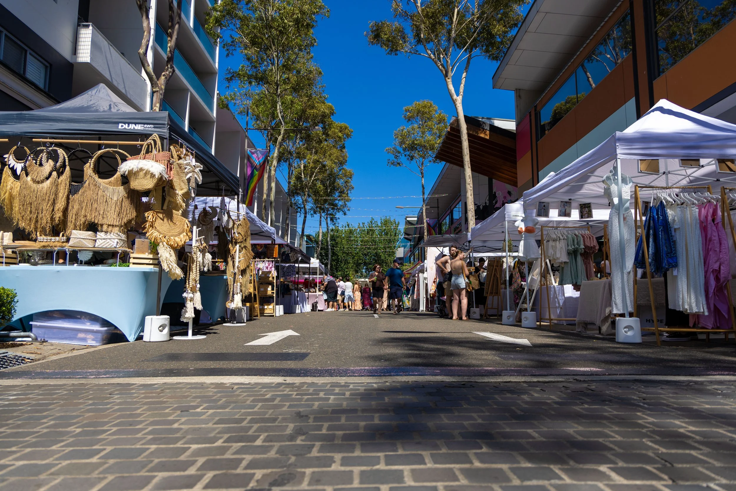 Outdoor street market with vendor booths selling clothing and accessories under white tents, people browsing, modern buildings, tall trees, a clear blue sky.