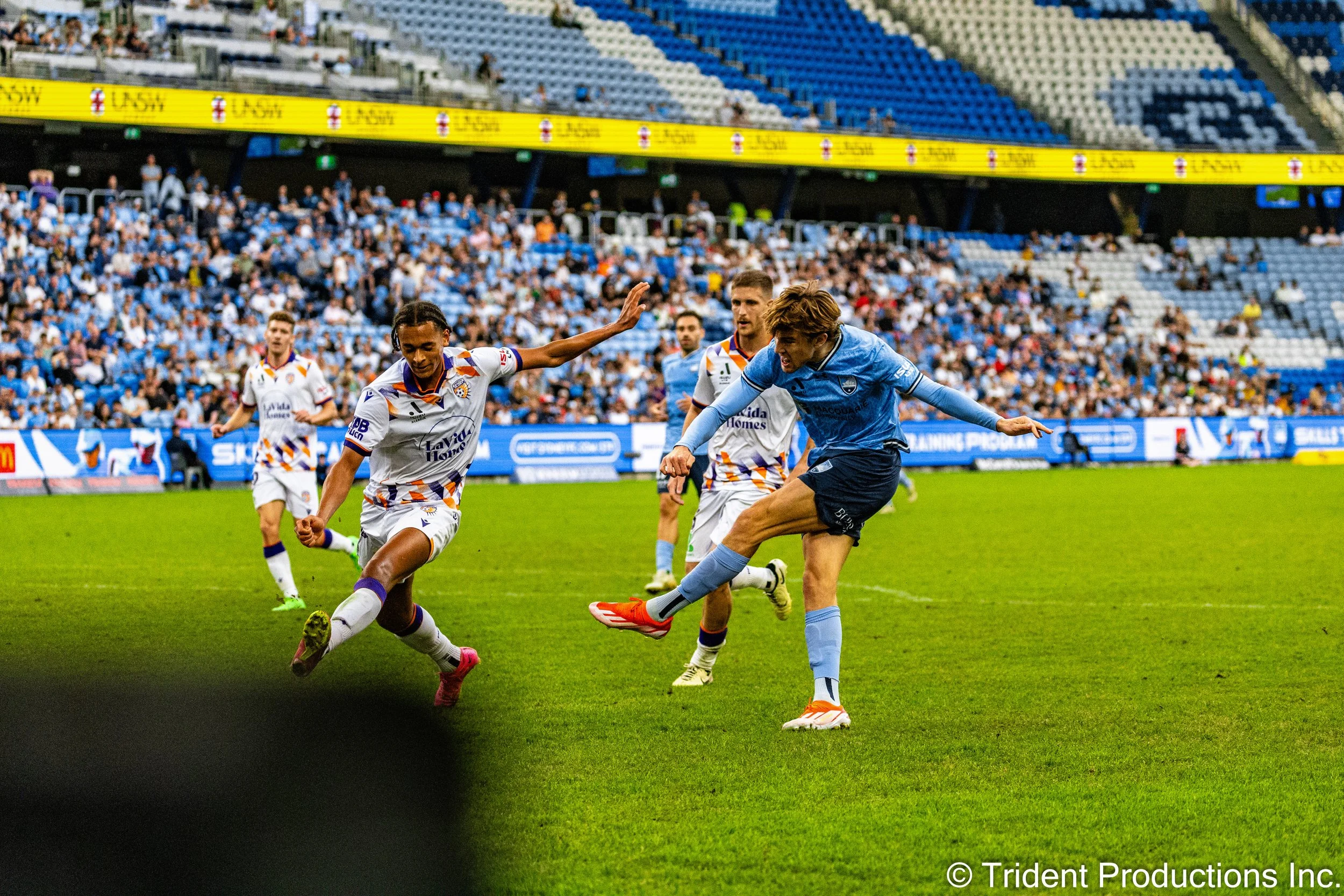 Soccer players in action during a match in a large stadium, with spectators in the background. One player in a light blue jersey is kicking the ball while opponents in white jerseys with orange and blue accents attempt to block or intercept.