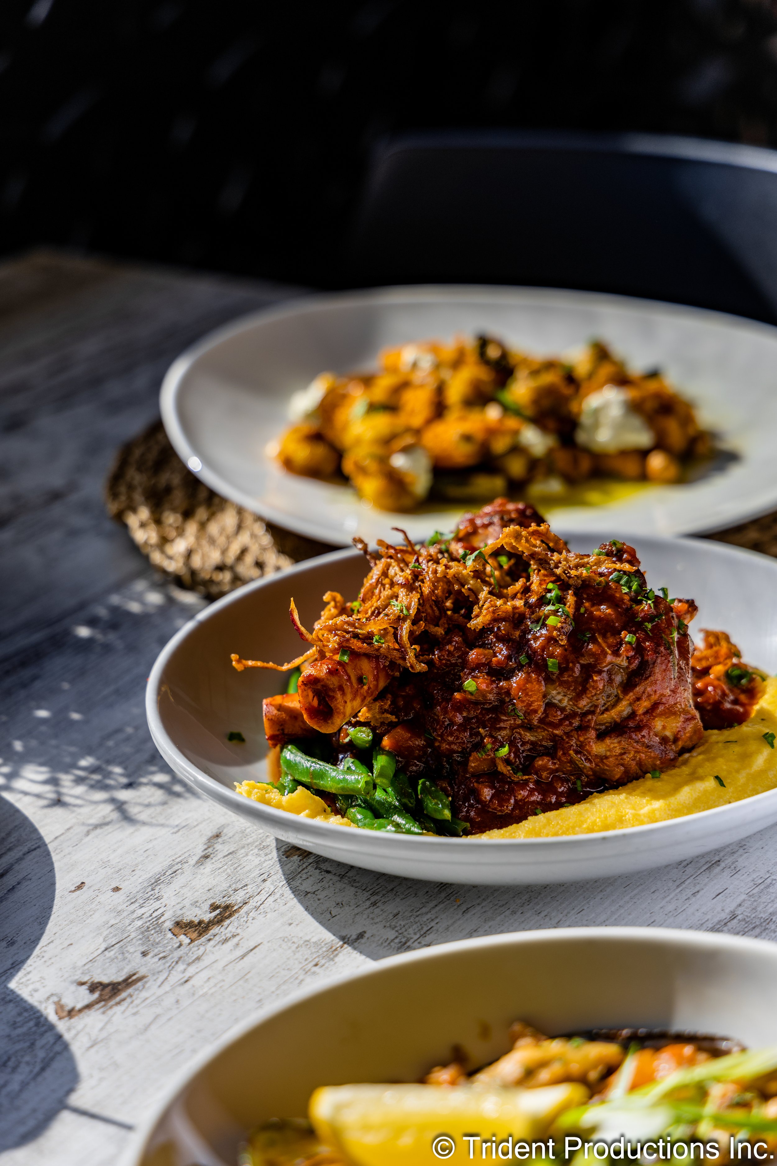 Three white plates containing different types of Indian cuisine dishes, including a plate with lamb, green beans, and yellow mashed potatoes in the foreground, a plate of spiced chicken or vegetable curry in the background, on a white wooden table with shadows and sunlight.