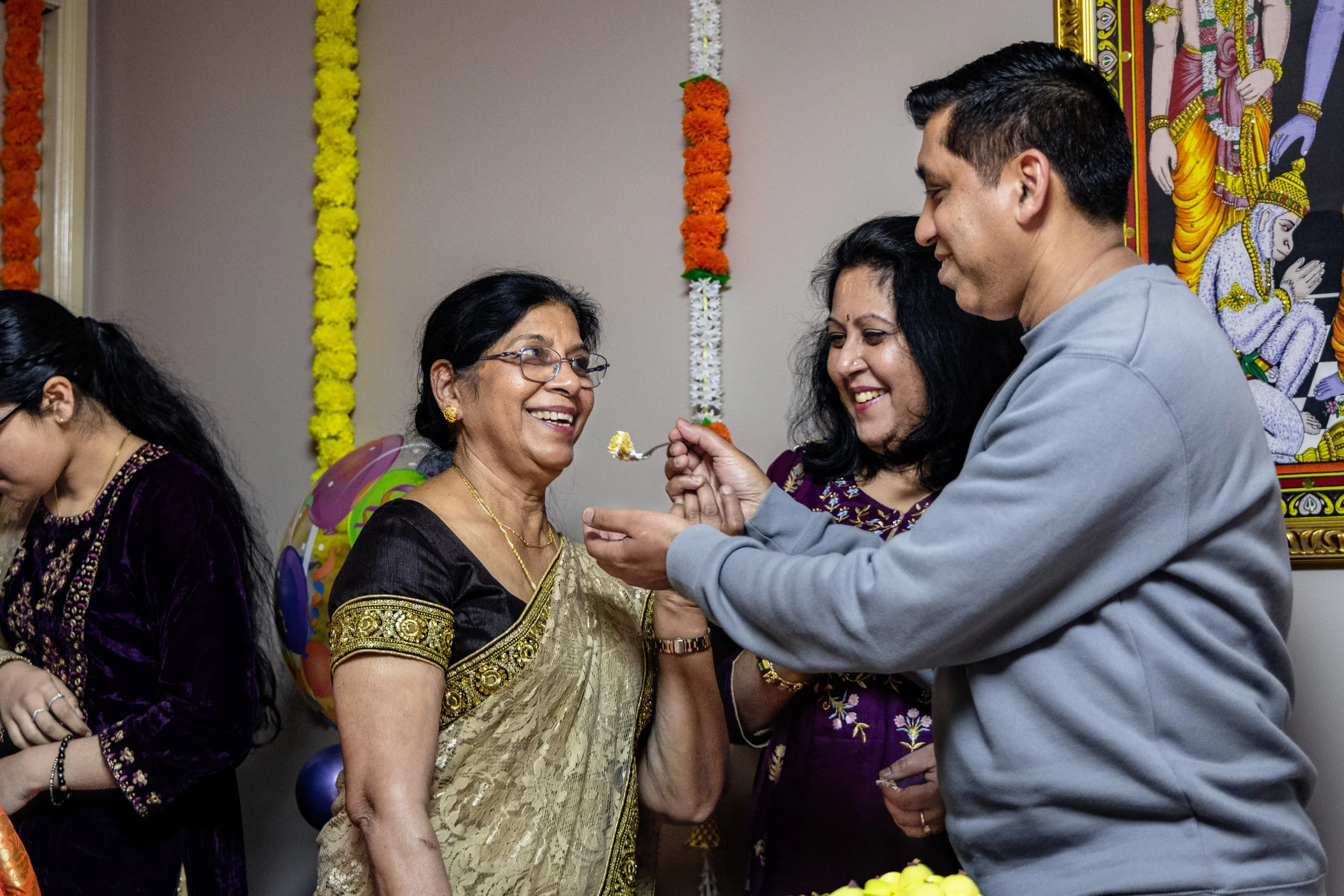 People celebrating during a traditional Indian ceremony, including a man feeding a woman. The woman is wearing a gold and black saree and glasses. The background has marigold flower decorations and a religious artwork.