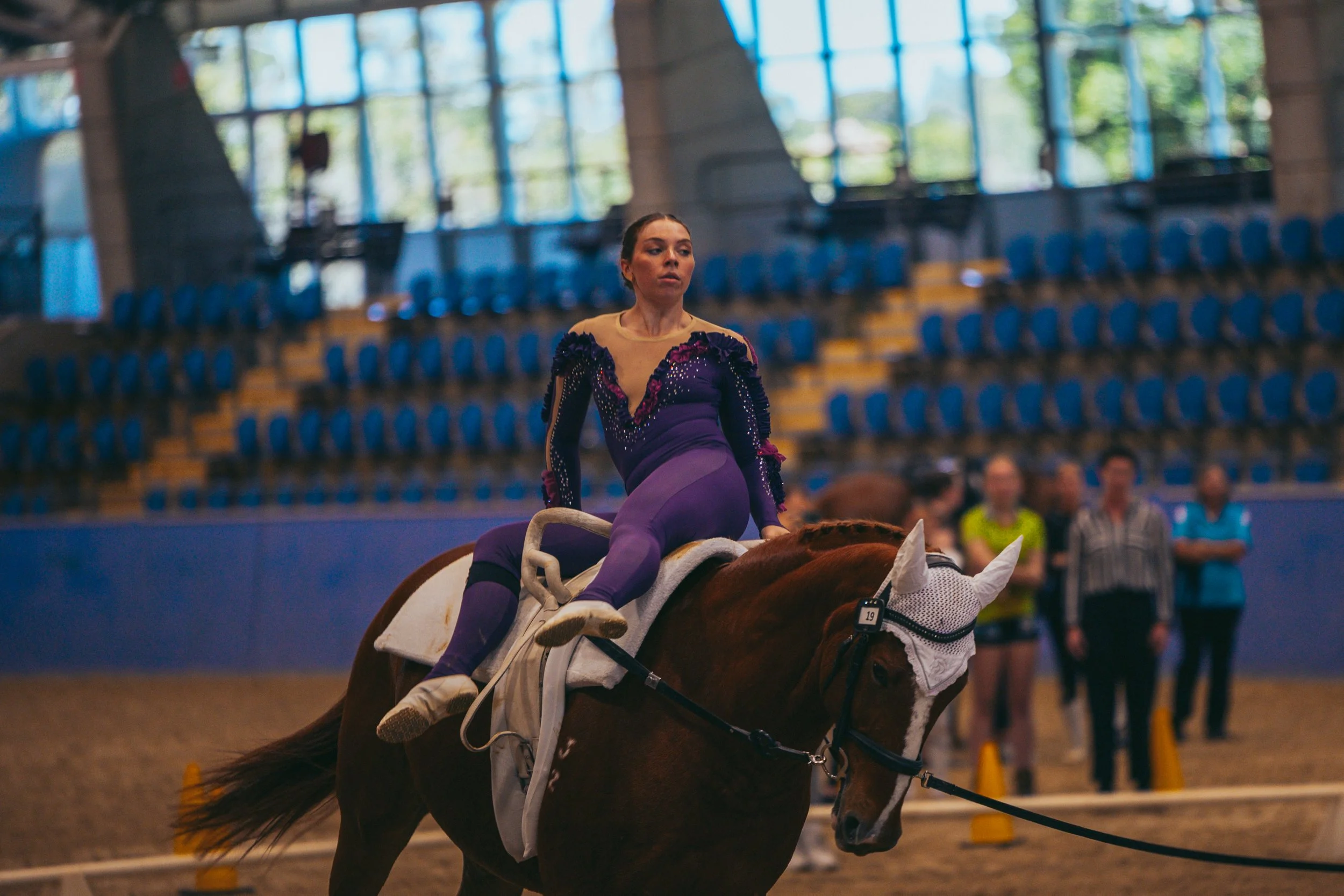 A female equestrian athlete in a purple and black costume riding a brown horse in an indoor riding arena, with a few spectators and officials in the background.