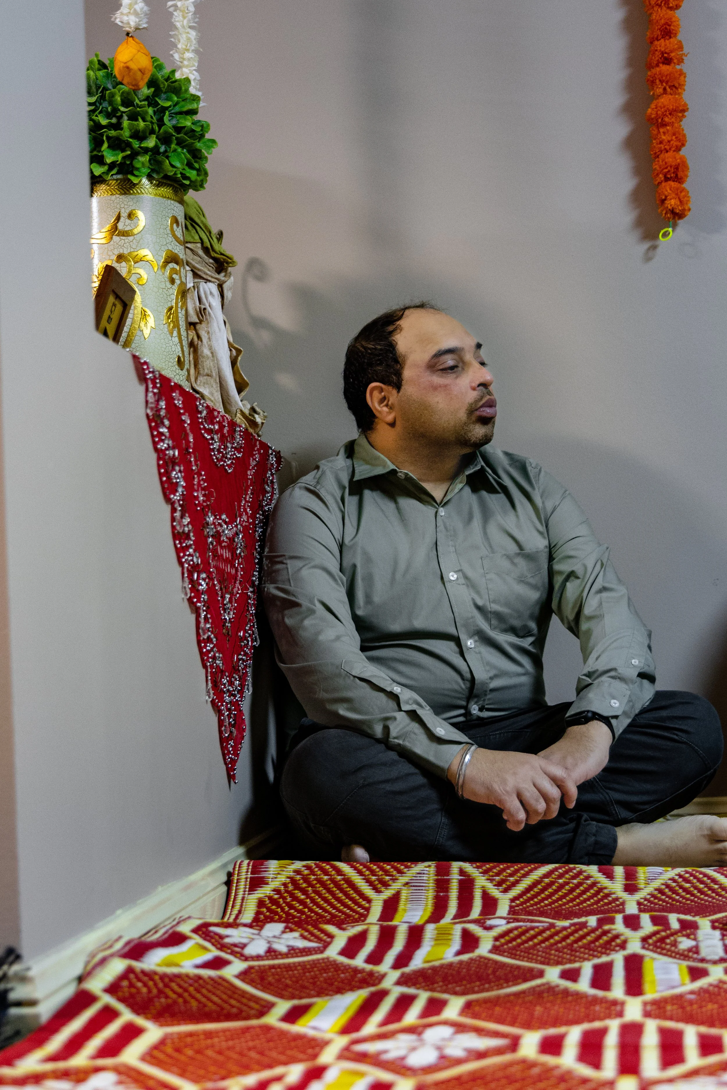 A man sitting on the floor with his eyes closed, surrounded by colorful decorations, including a red and yellow patterned cloth on the floor, a floral garland hanging, and a decorative plant in a pot.