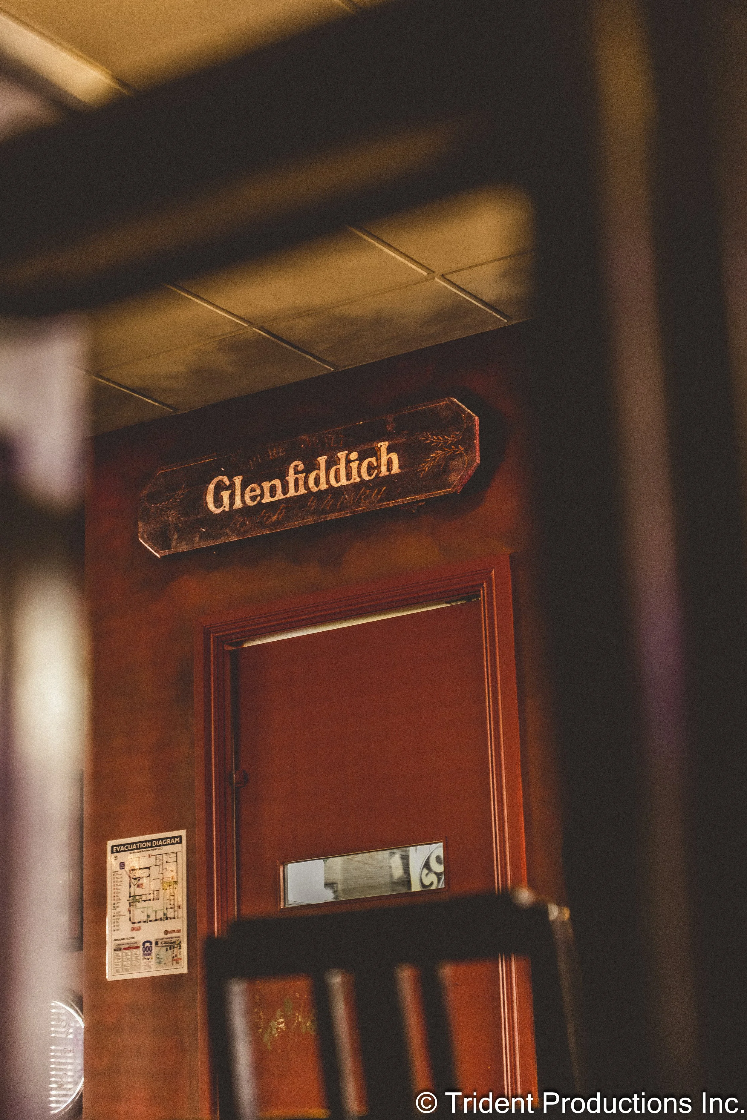 A bar scene showing a door with a small window, a fire evacuation diagram on the wall, and a sign with the name 'Glenfiddich' above the door.