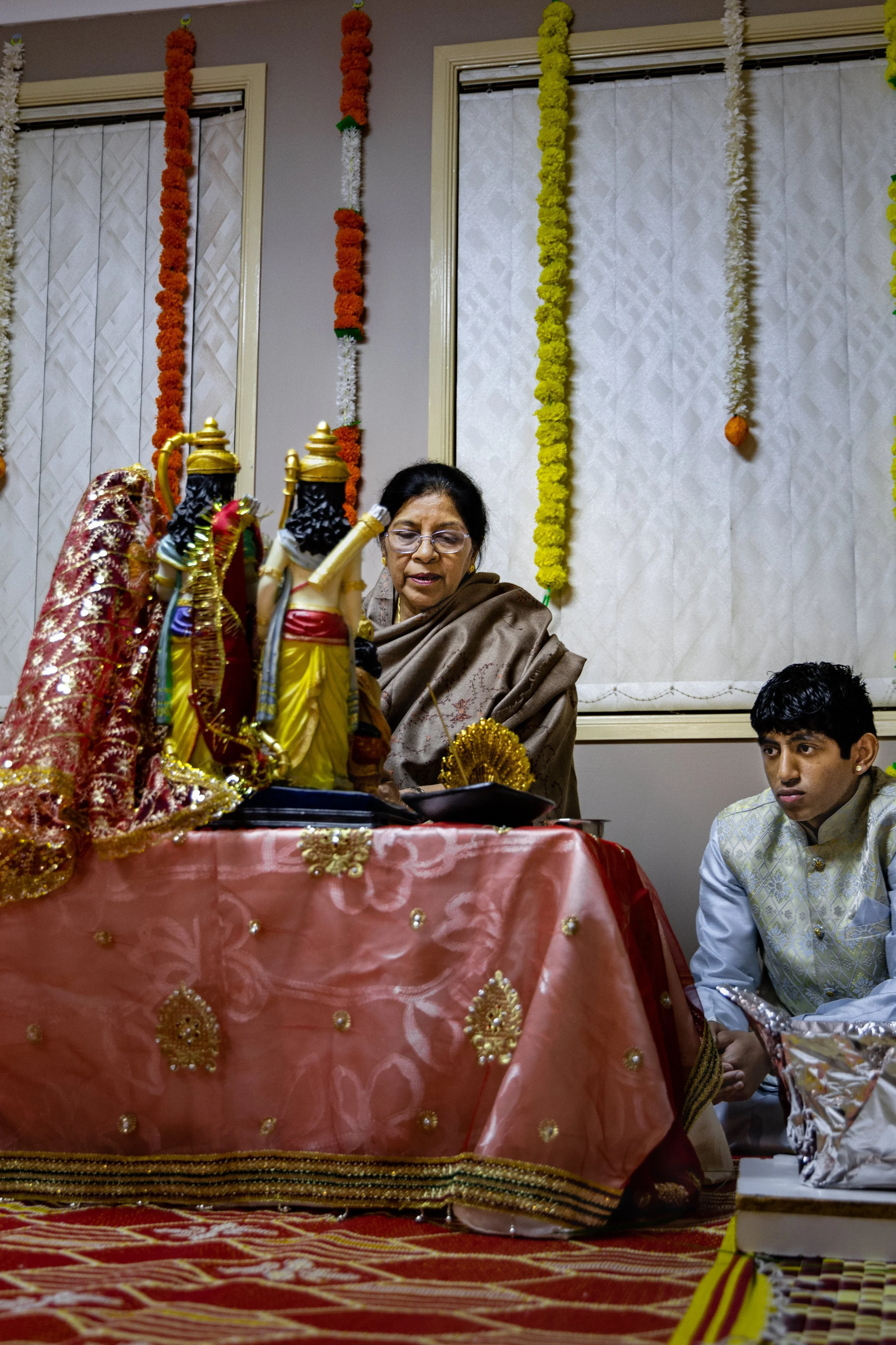 An Indian woman and a young man are participating in a religious or cultural ceremony, seated in front of decorated idols and offerings, with colorful garlands hanging on the wall behind them.