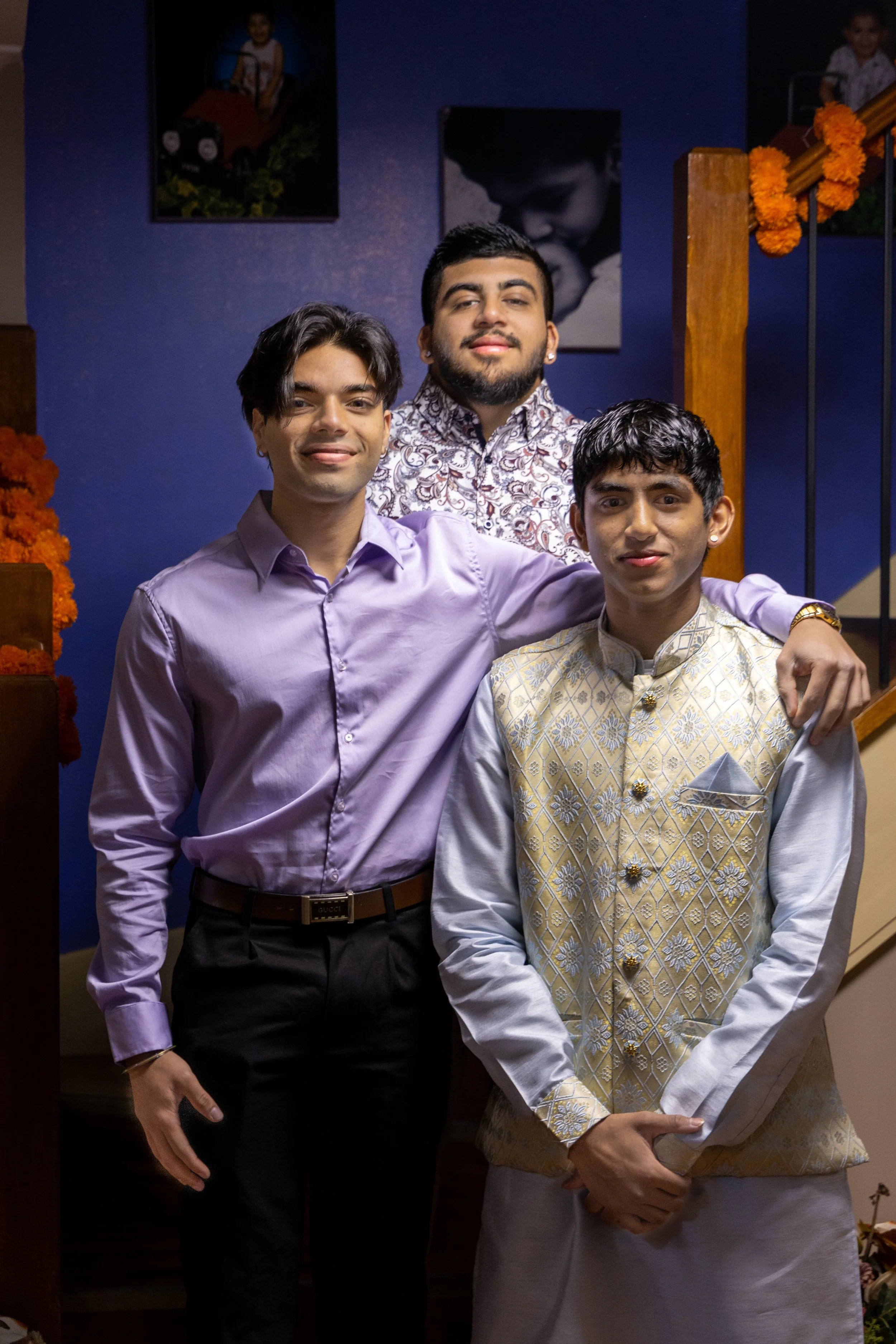 Three young men dressed in formal and traditional Indian attire at a celebration or gathering in a decorated indoor setting.