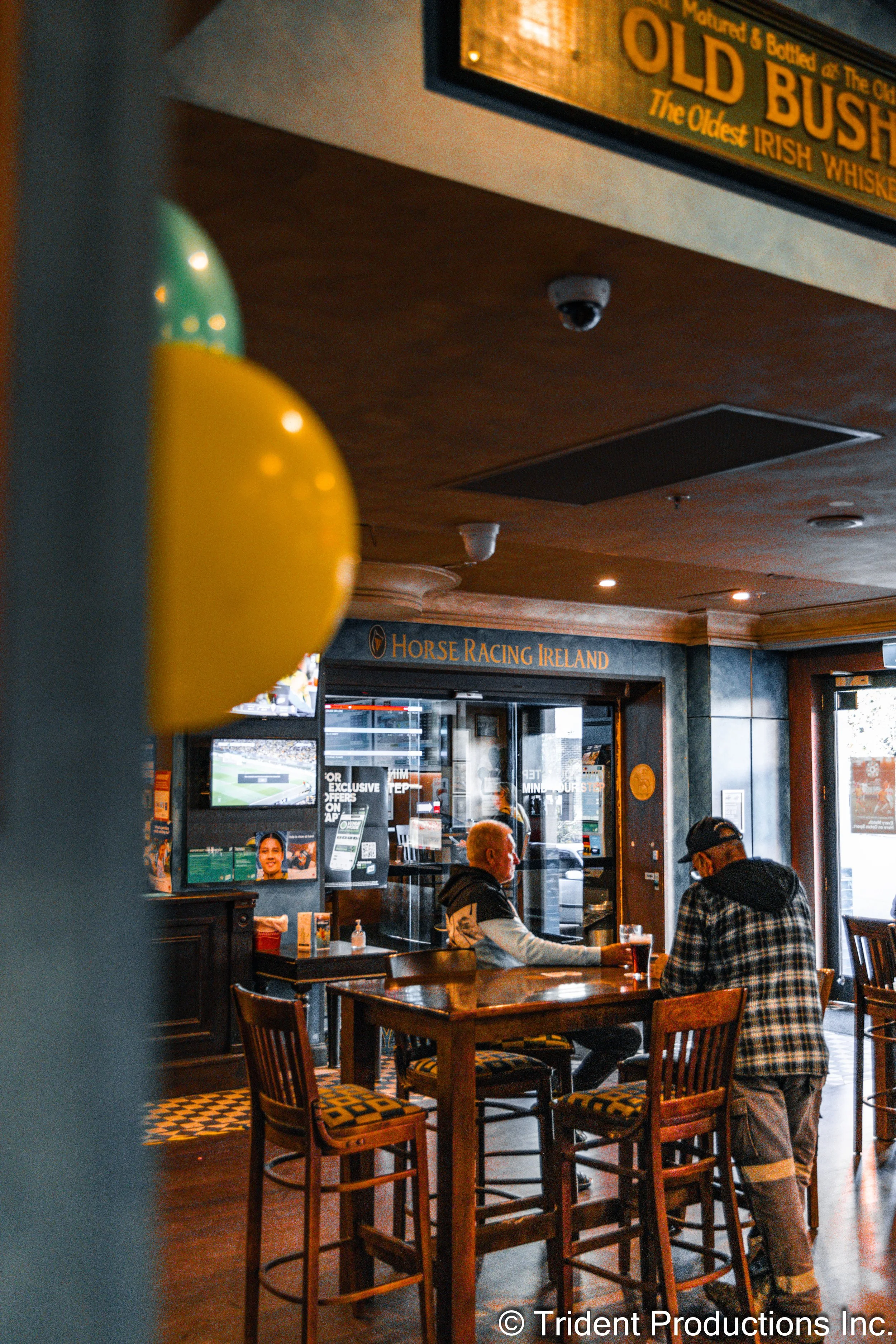 Two men sitting at a wooden high-top table in a pub, with a TV and sports betting machines in the background, and balloons partially visible in the foreground.