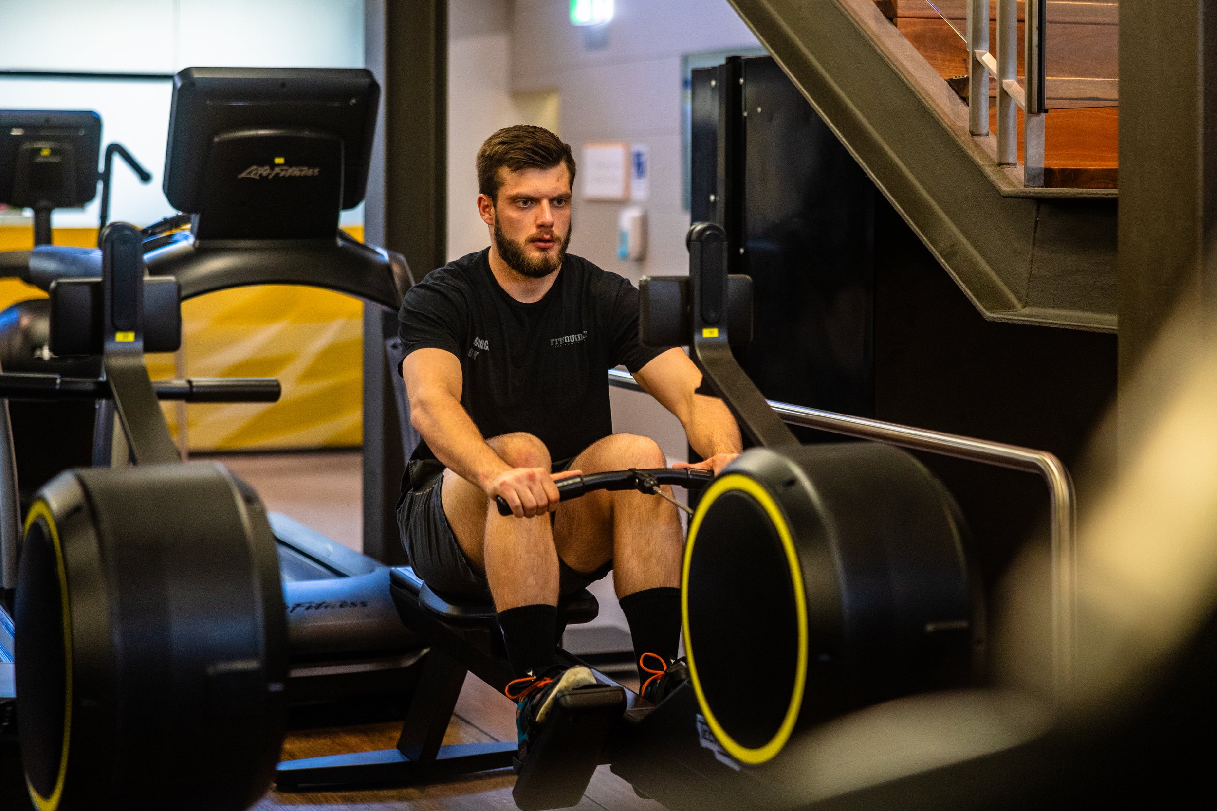 A man with a beard wearing a black t-shirt and black shorts working out on a rowing machine in a gym.