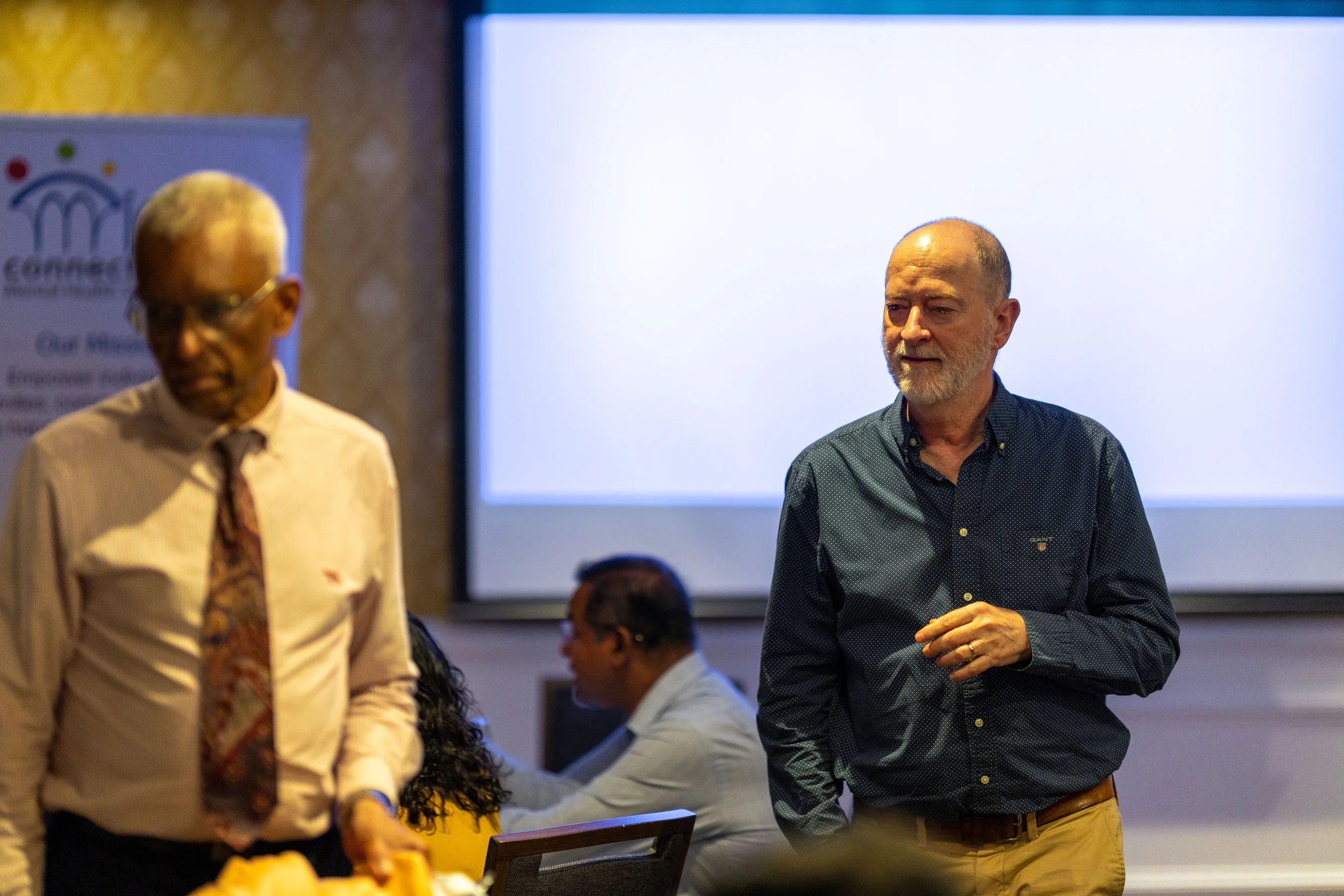 Two men are standing in a conference room, one in the foreground wearing a navy blue shirt and tan pants, and the other in the background wearing glasses, a light-colored shirt, and a patterned tie. There are people sitting and working at a table nea