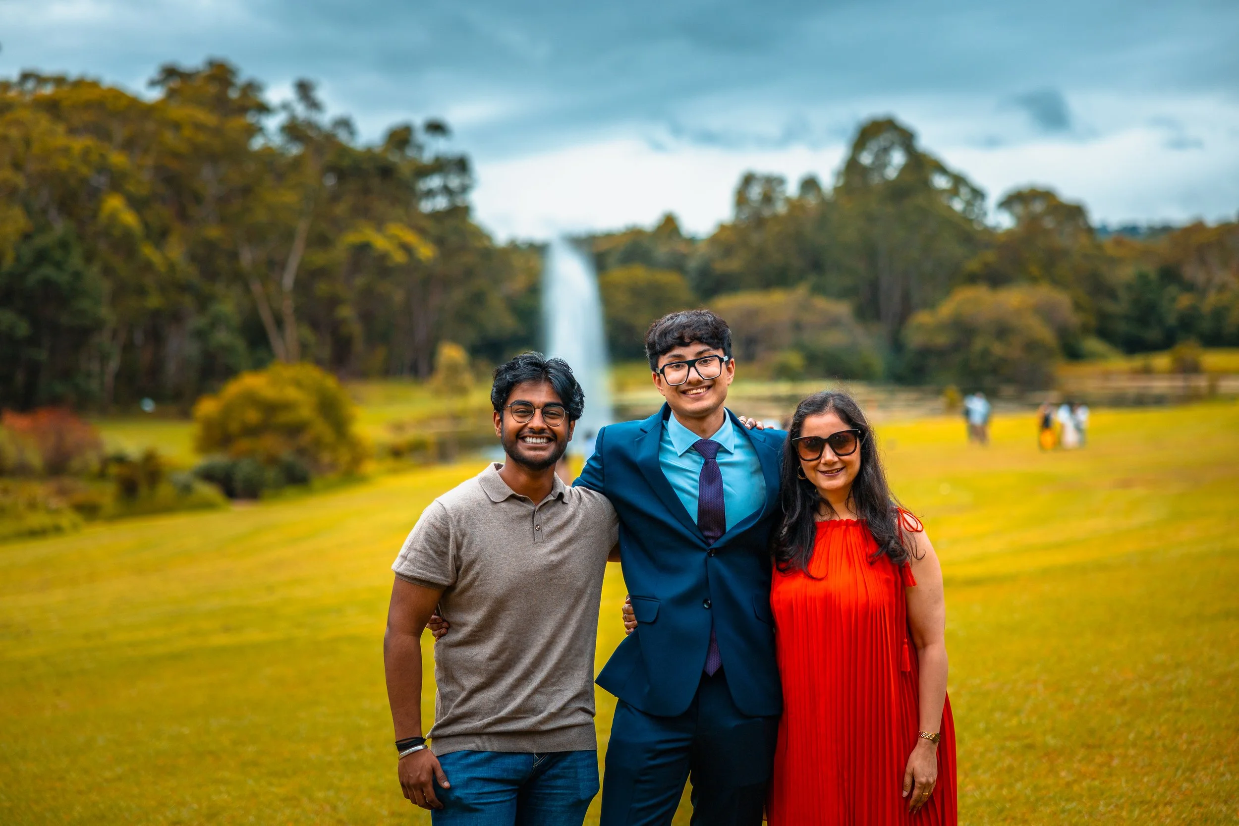 Three people smiling in a park with a waterfall and trees in the background.