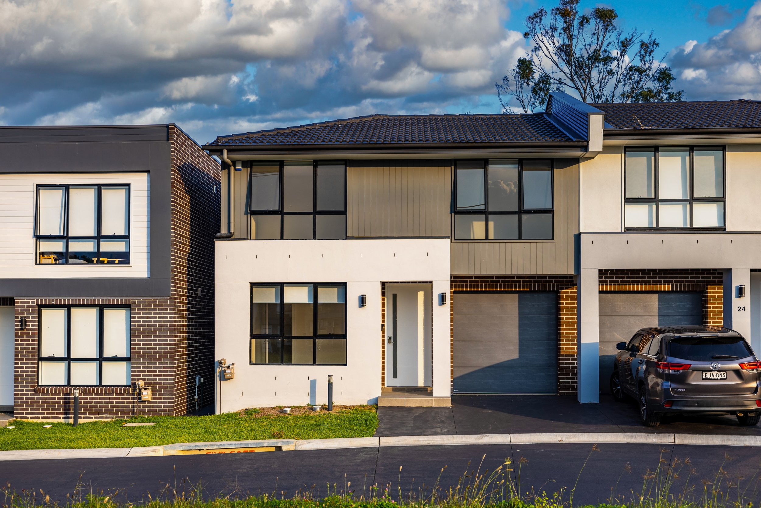 Modern two-story rowhouse with gray and beige exterior, large front window, black front door, gray garage door, and black car parked in driveway under a partly cloudy sky.