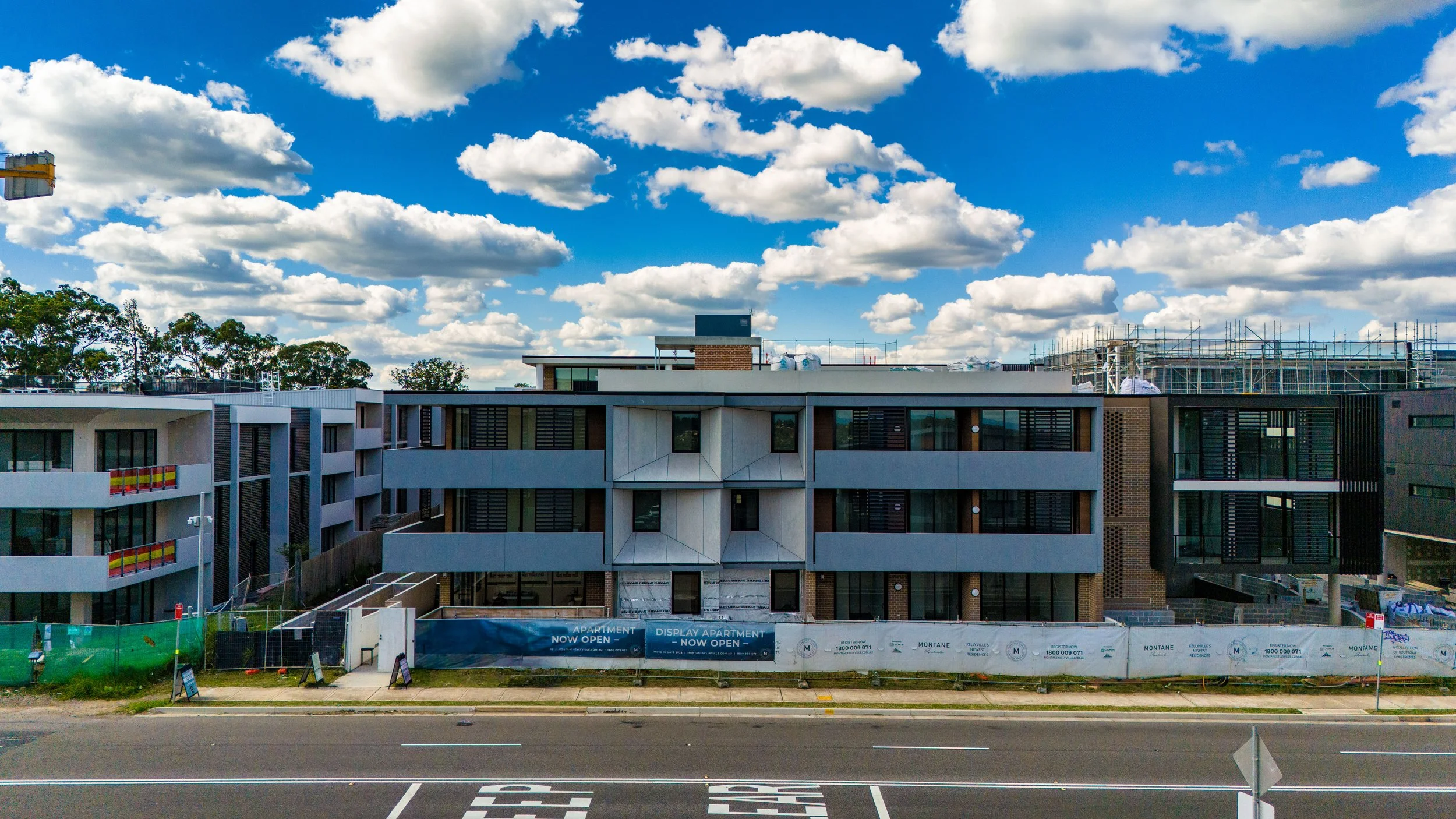 Modern multi-story apartment building under construction with a street in the foreground and a bright blue sky with scattered clouds in the background.