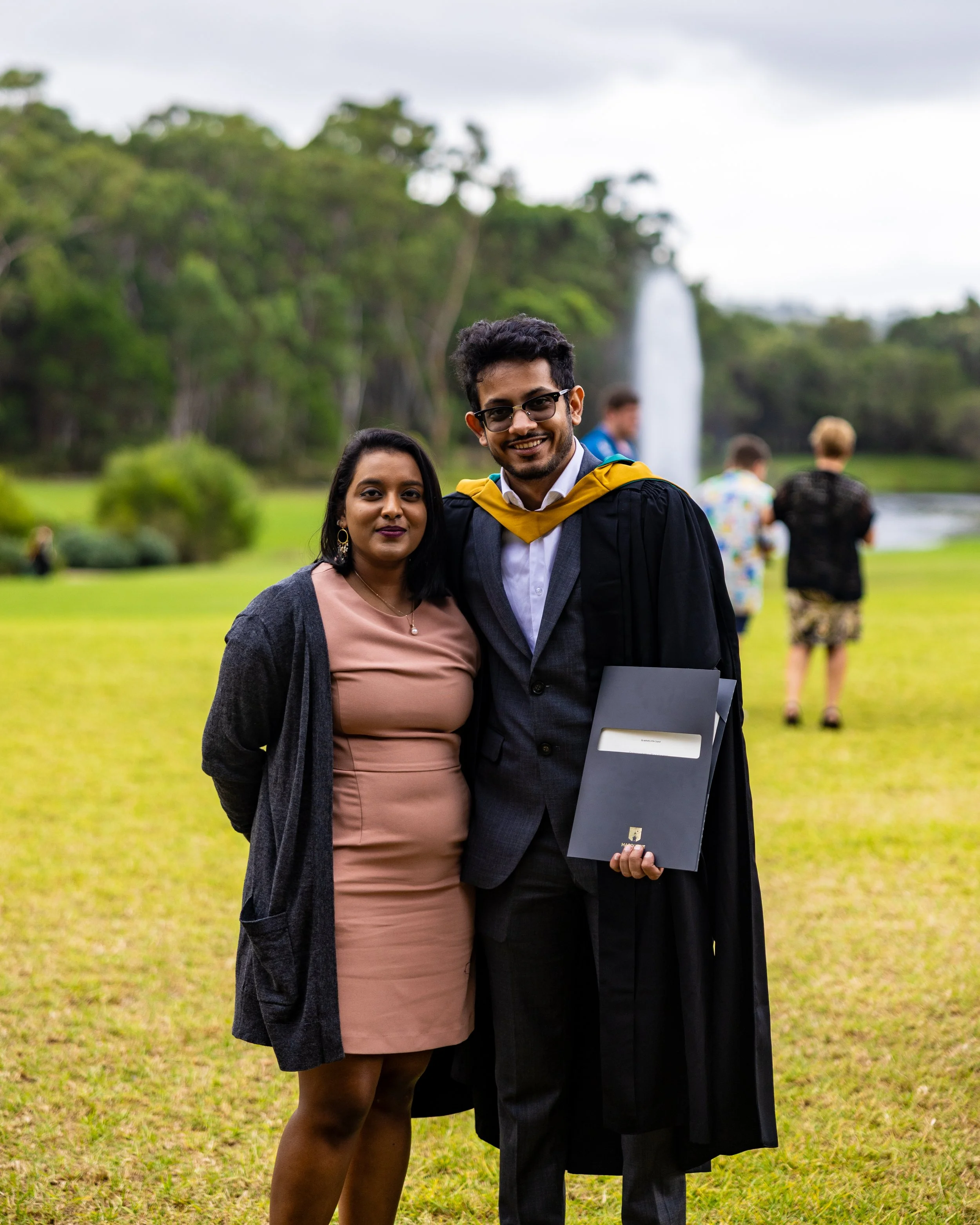 A young man in a graduation gown and cap holding a folder, standing with a woman on a grassy field with trees and a fountain in the background.