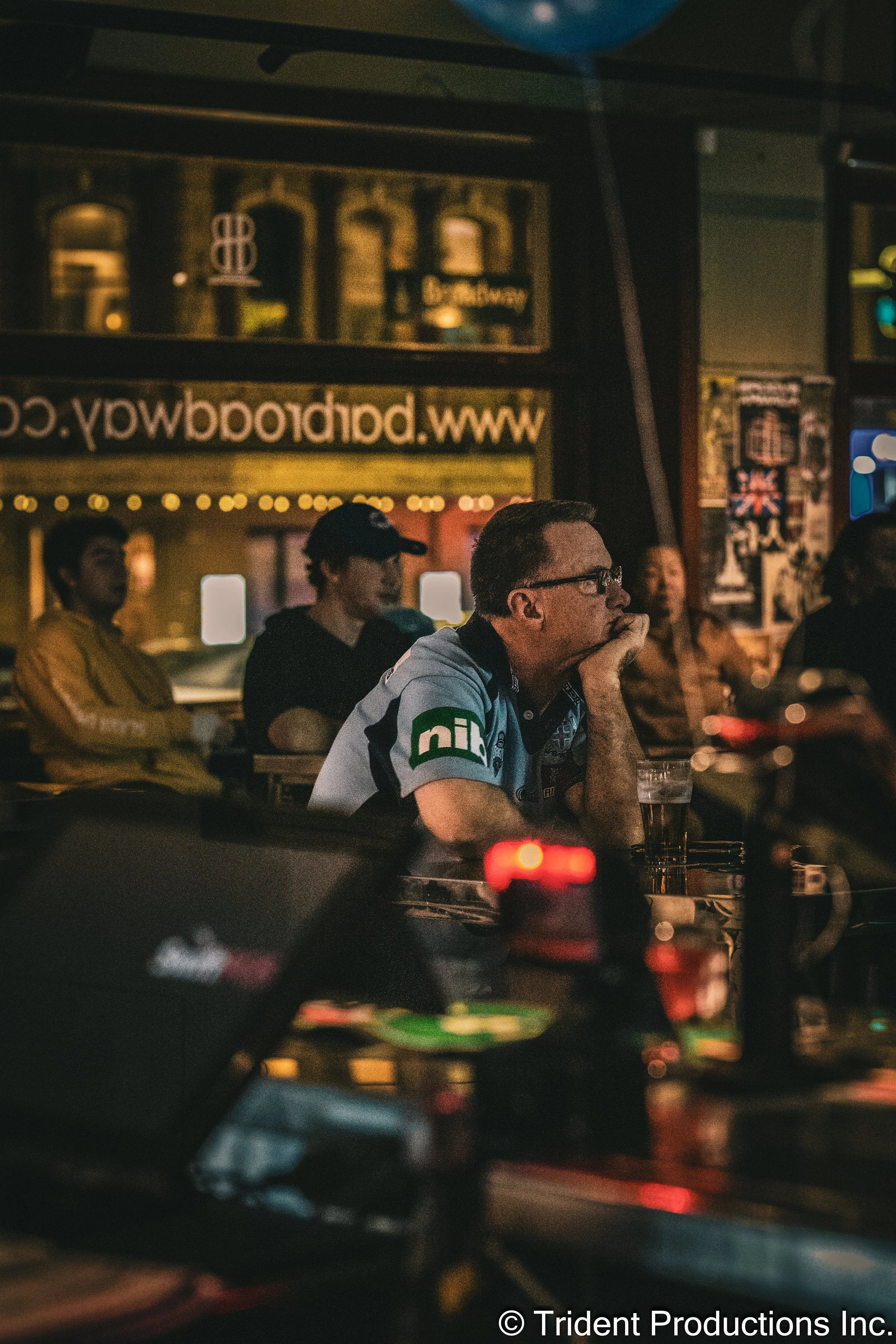 People sitting at a bar or pub, watching something intently with a man in a racing jersey in the foreground and others in the background.