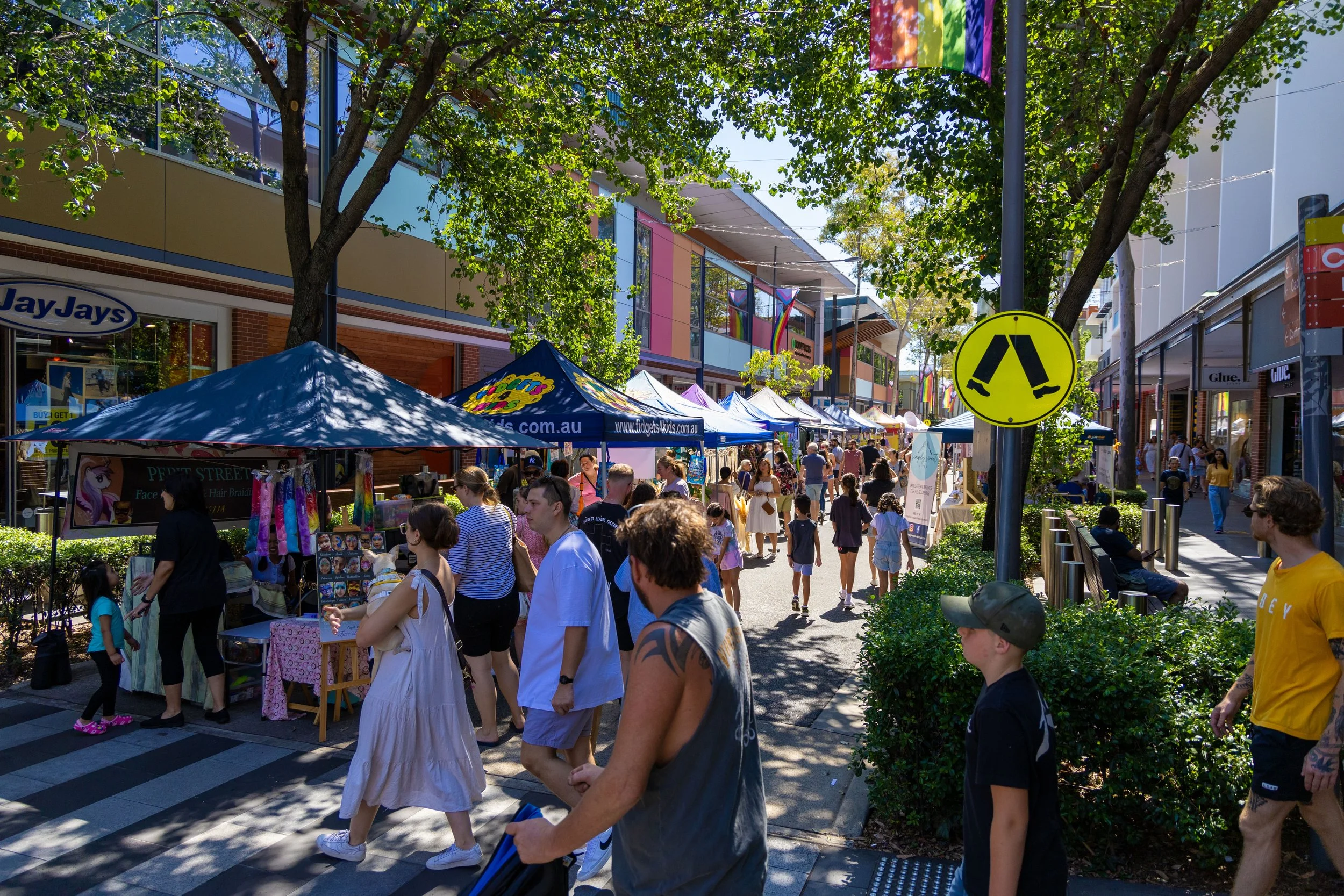A busy outdoor shopping street with vendors under tents, people walking and browsing, and trees providing shade. A bright yellow pedestrian crossing sign is visible.