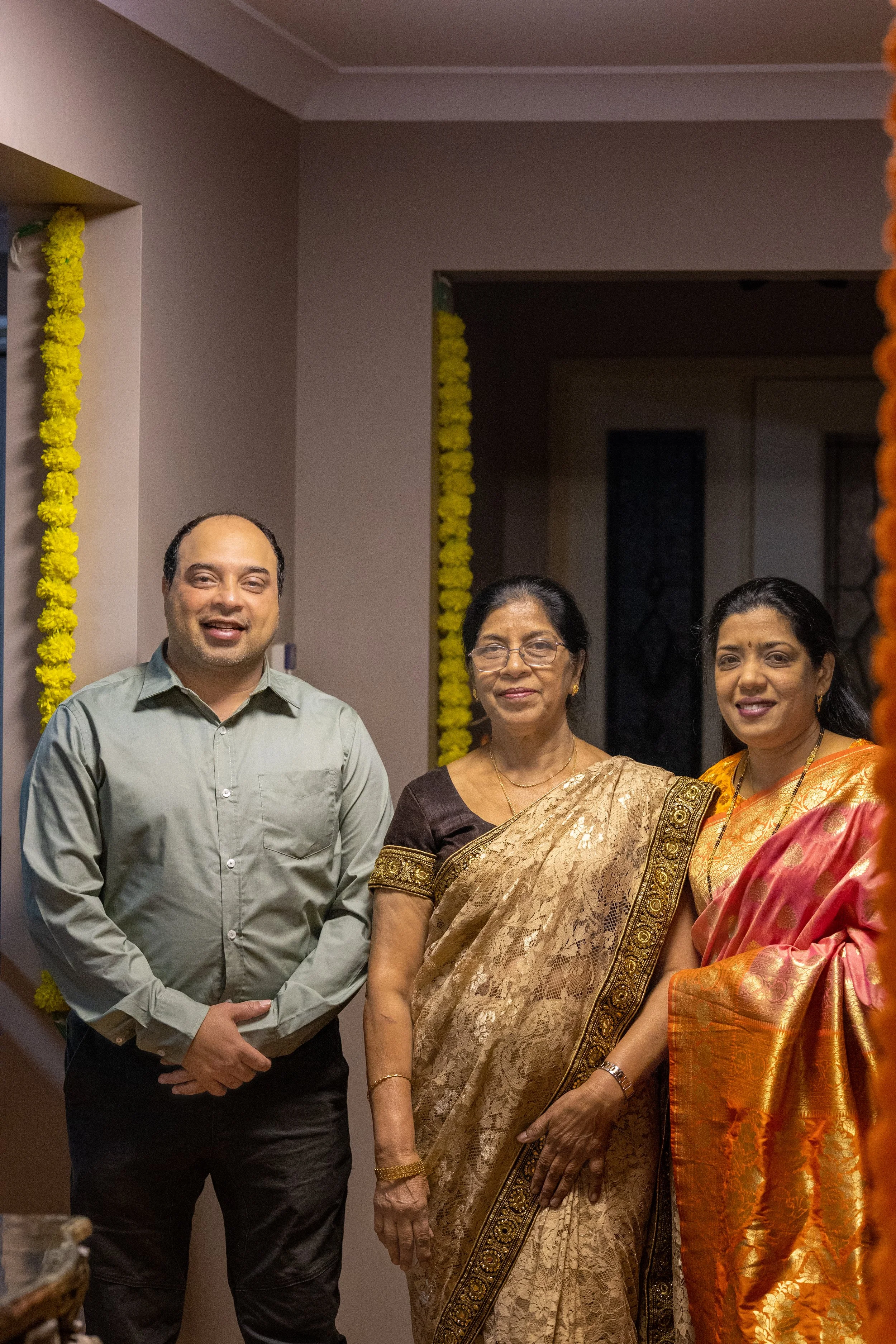 Three people standing together, two women wearing traditional Indian sarees and a man in formal attire, inside a decorated room with yellow flower garlands.
