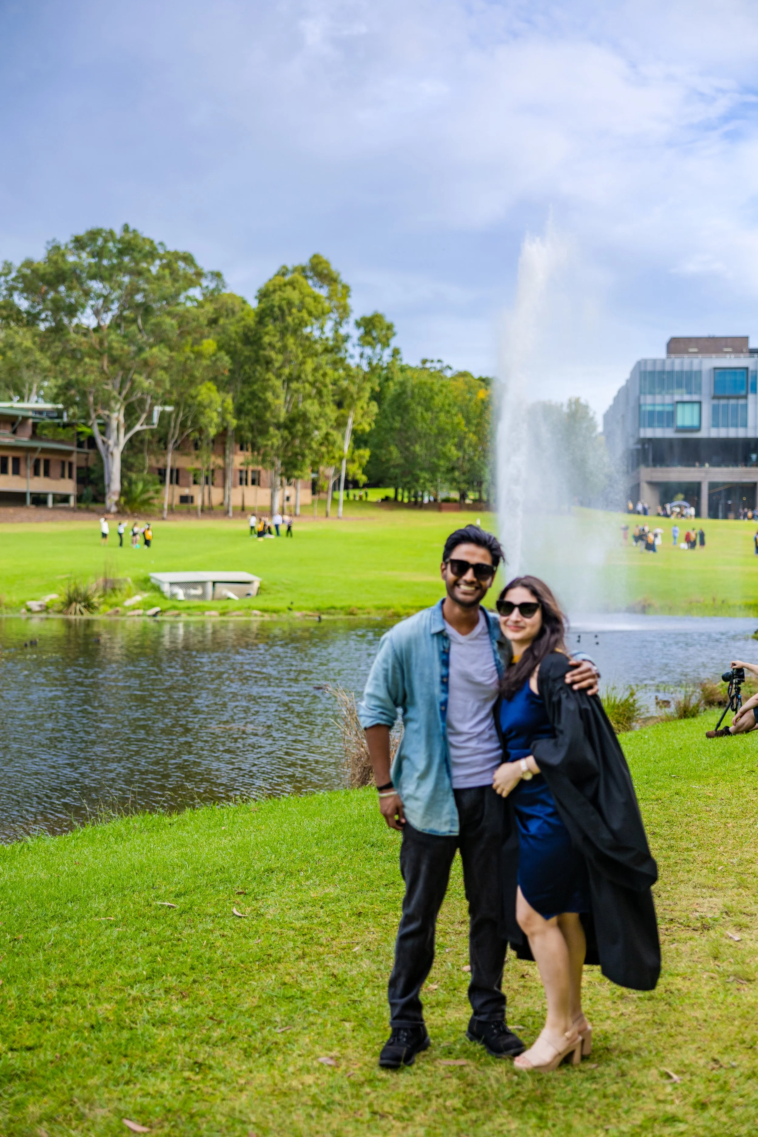 A smiling couple standing on grass by a pond with a fountain, surrounded by trees and buildings, during daytime.
