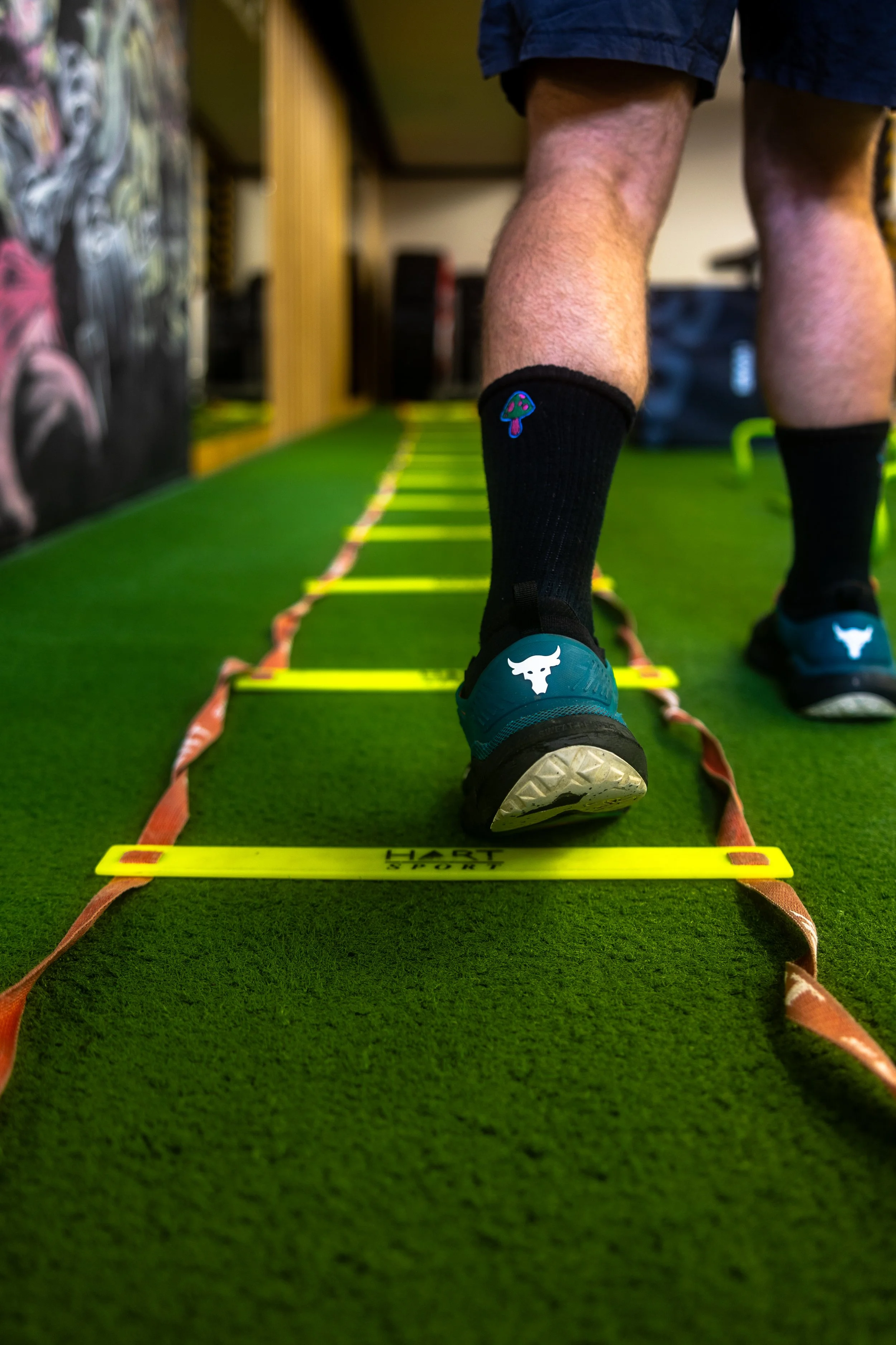 Close-up of a person's legs and feet as they walk on a gym agility ladder on green turf, wearing black socks with a colorful mushroom logo and blue athletic shoes with a white bull head logo.