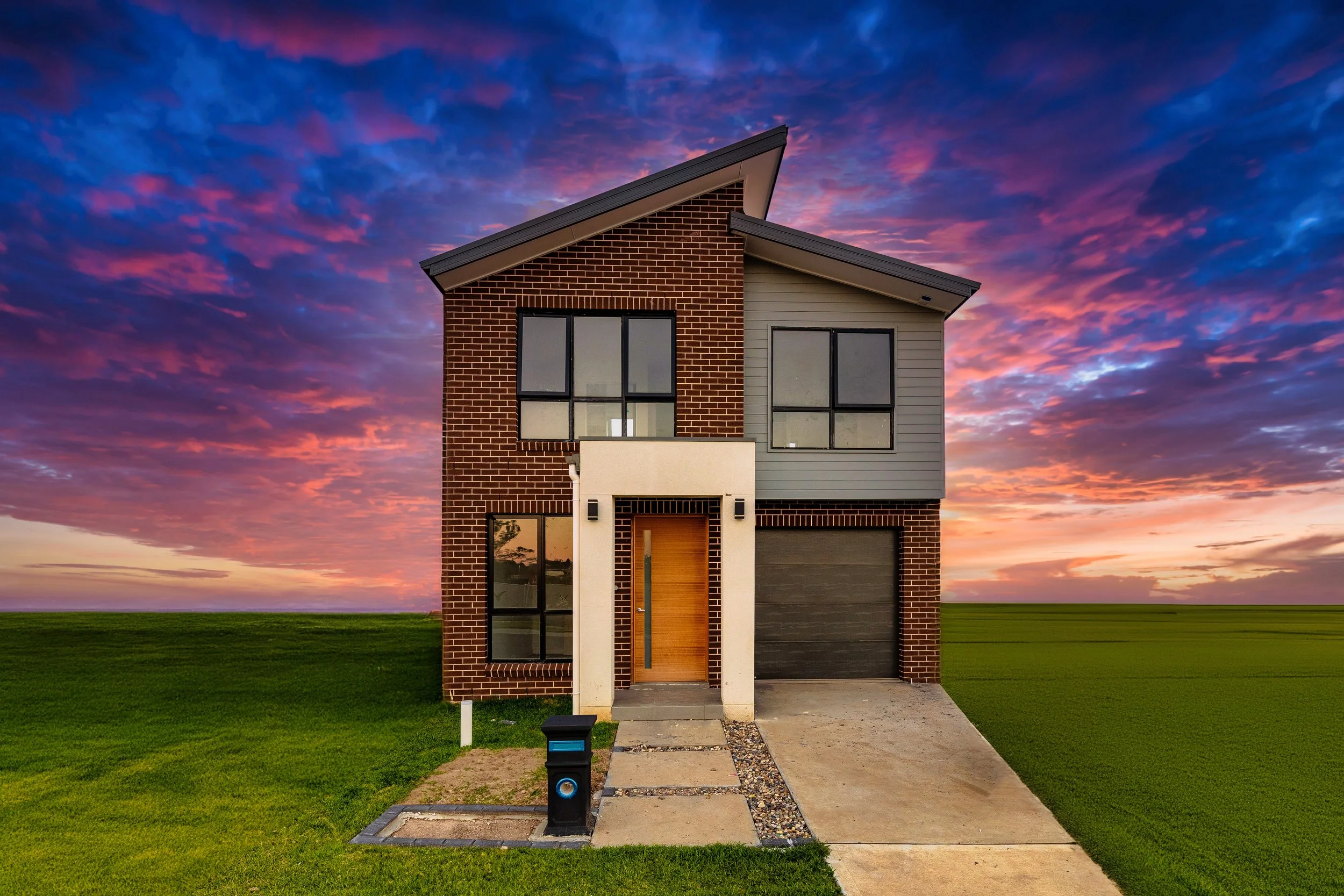 Modern two-story house with brick and gray siding exterior, wooden front door, and large windows, set on a landscaped lawn at sunset with colorful sky.