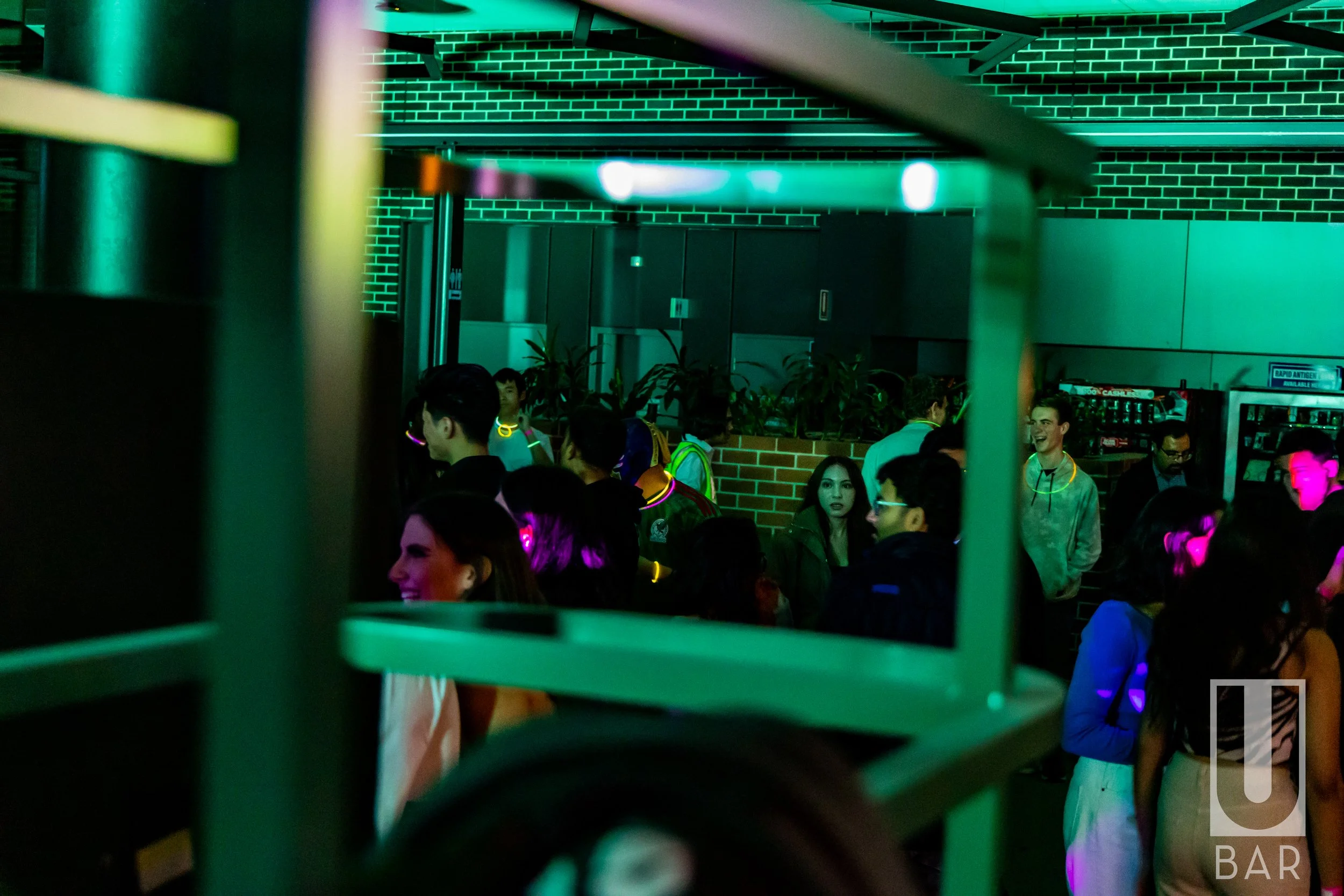 People socializing at a bar or club with green lighting, neon glow necklaces, and a brick wall in the background.