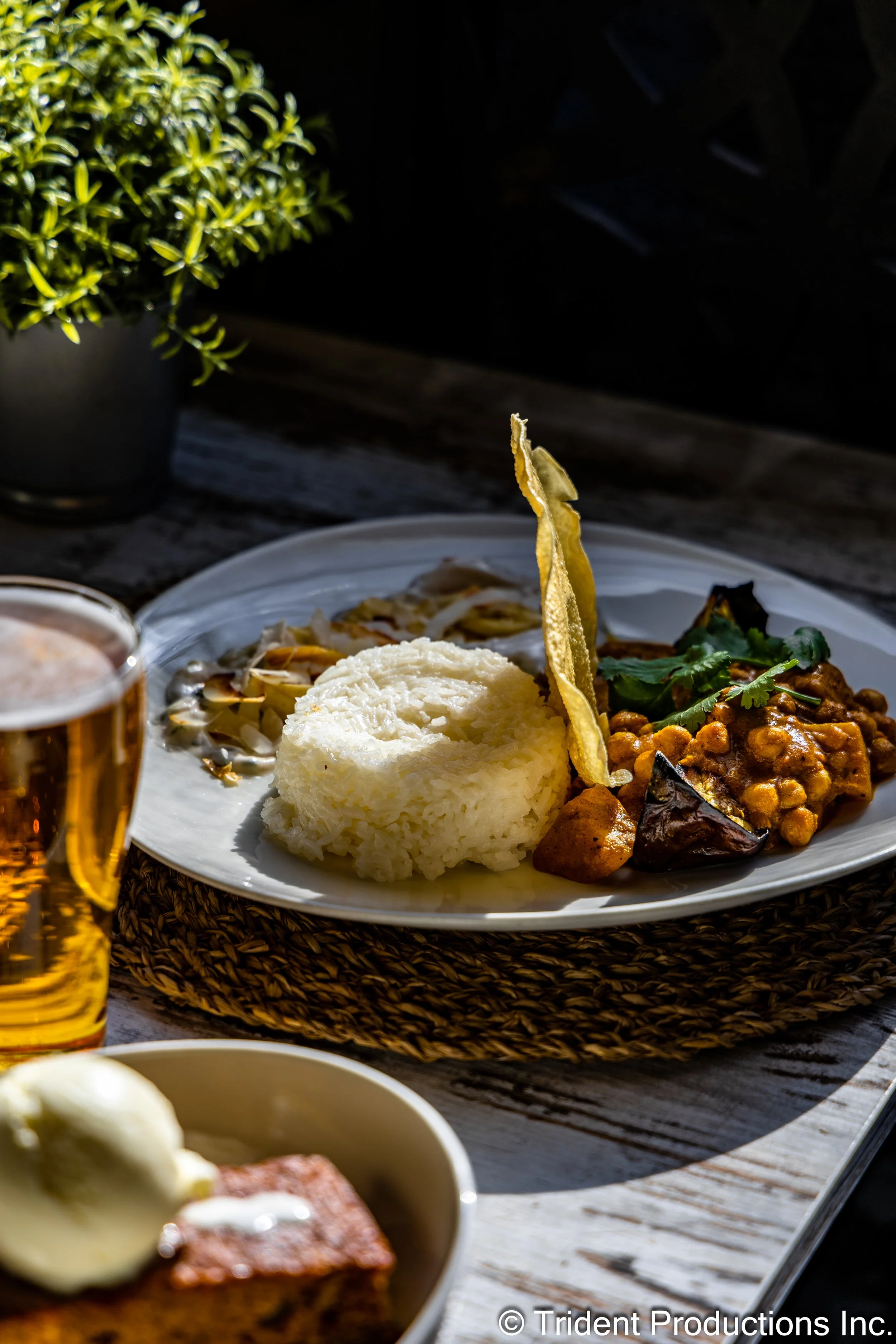 A plate of Indian curry with chickpeas, mashed potatoes, and roasted vegetables, garnished with fresh cilantro, served on a woven placemat with a splash of sunlight.