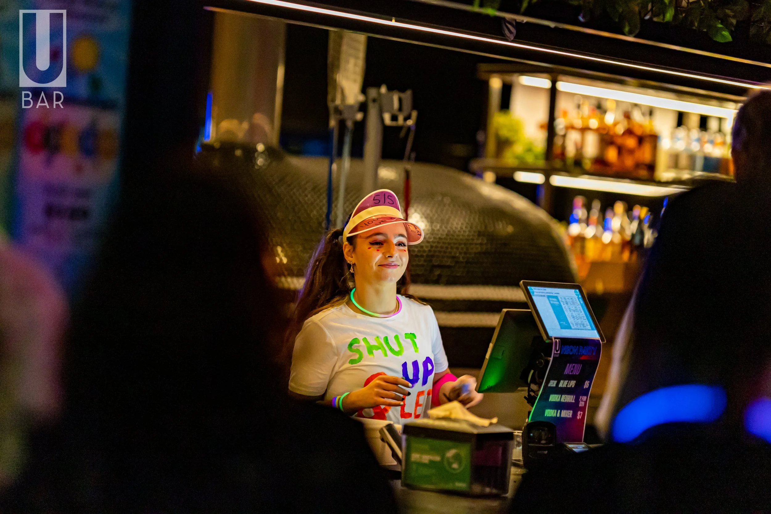 A young woman working at a bar, wearing a colorful shirt with the words 'SHUT UP' in bright letters, a plastic visor with '515' written on it, and glow necklaces, in a dimly lit environment with a neon-lit bar and people around.