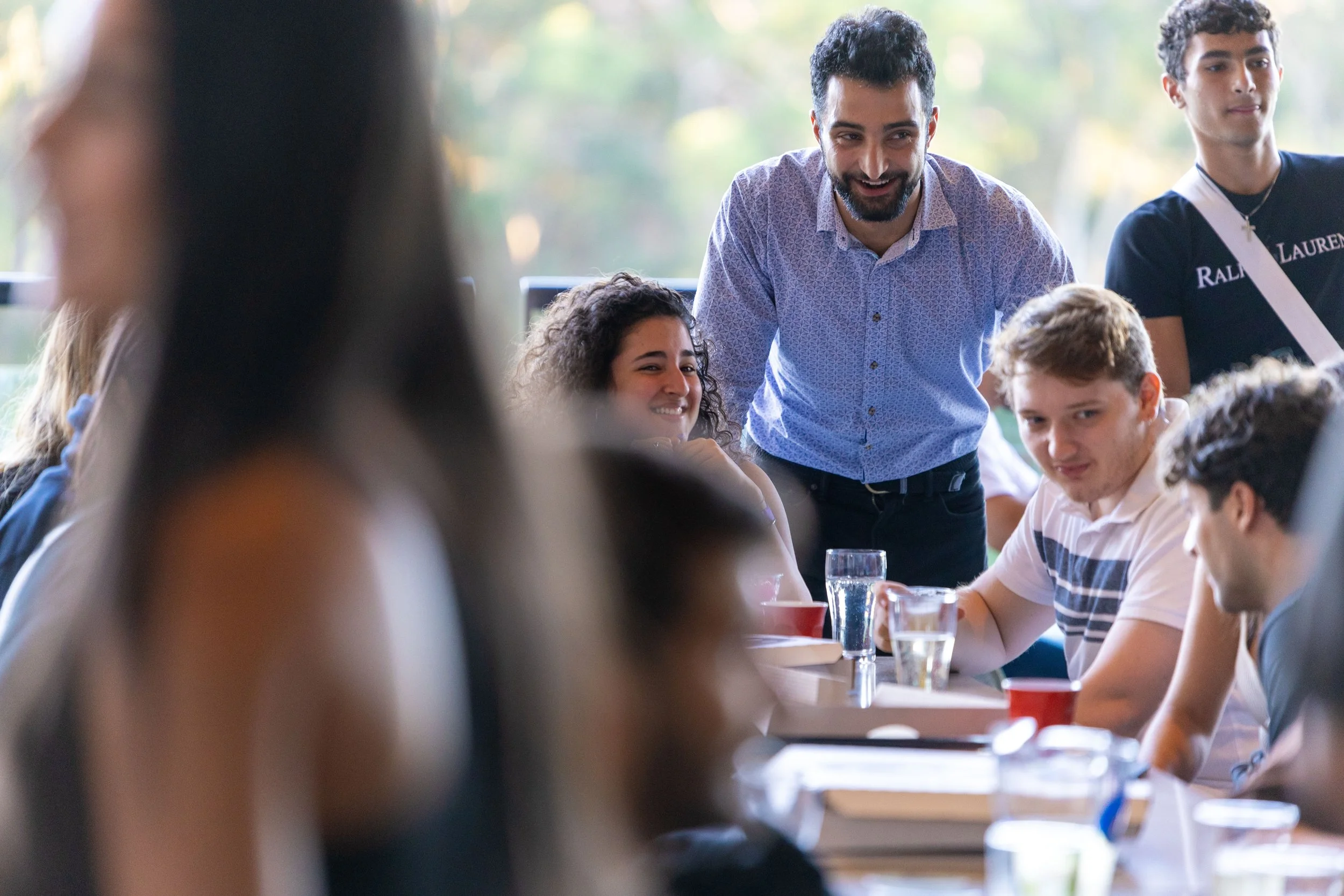 A group of diverse young people and a man are gathered around a table, smiling and engaging in conversation, with drinks and books on the table, in a bright, outdoor setting.