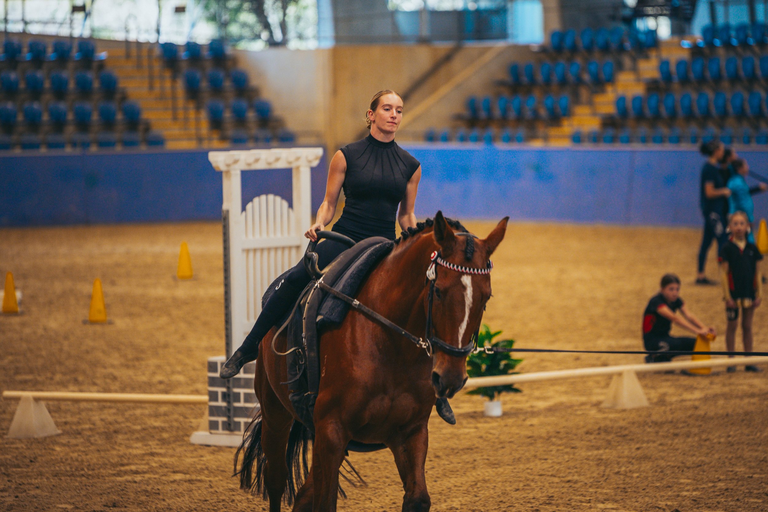 A woman riding a brown horse inside an indoor arena, with blue and yellow seating in the background and children playing.