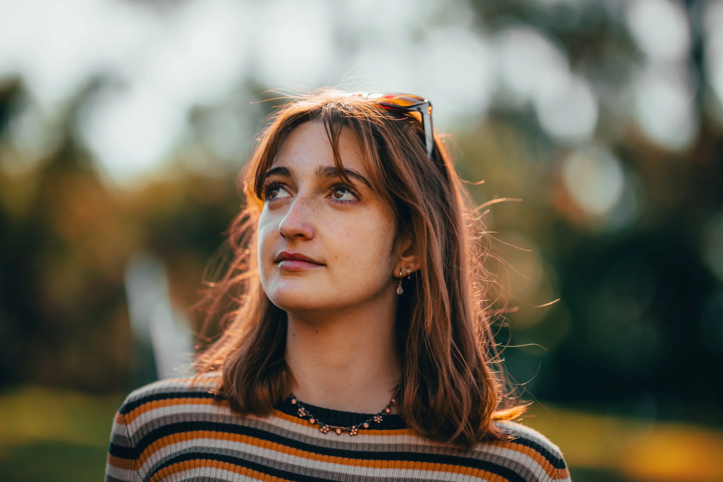 Young woman outdoors with sunglasses on her head, looking up with a slight smile, wearing a striped shirt and jewelry, in natural light