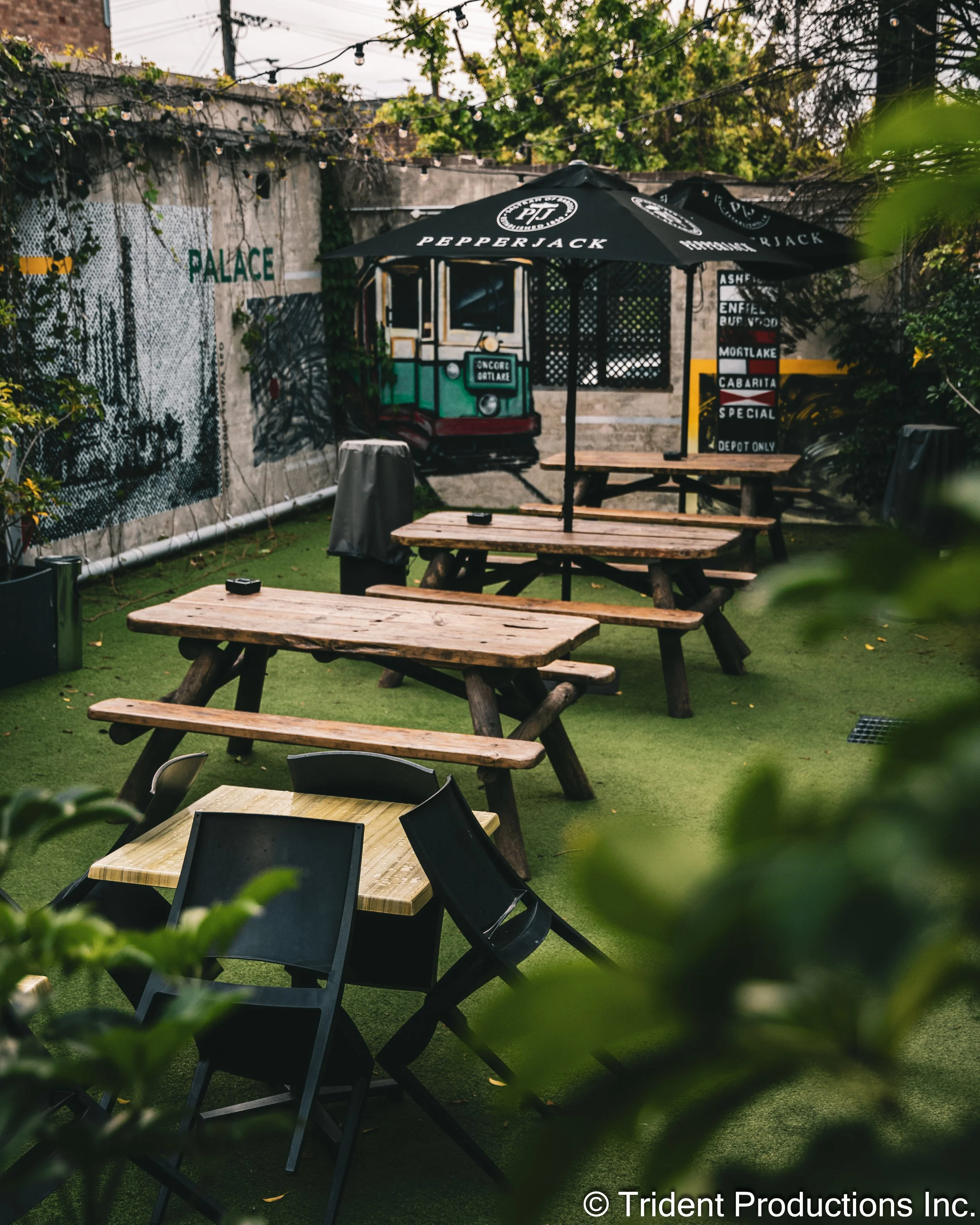 Outdoor patio with wooden picnic tables, black chairs, a large umbrella with Pepperjack branding, a mural of a streetcar, potted plants, and string lights.